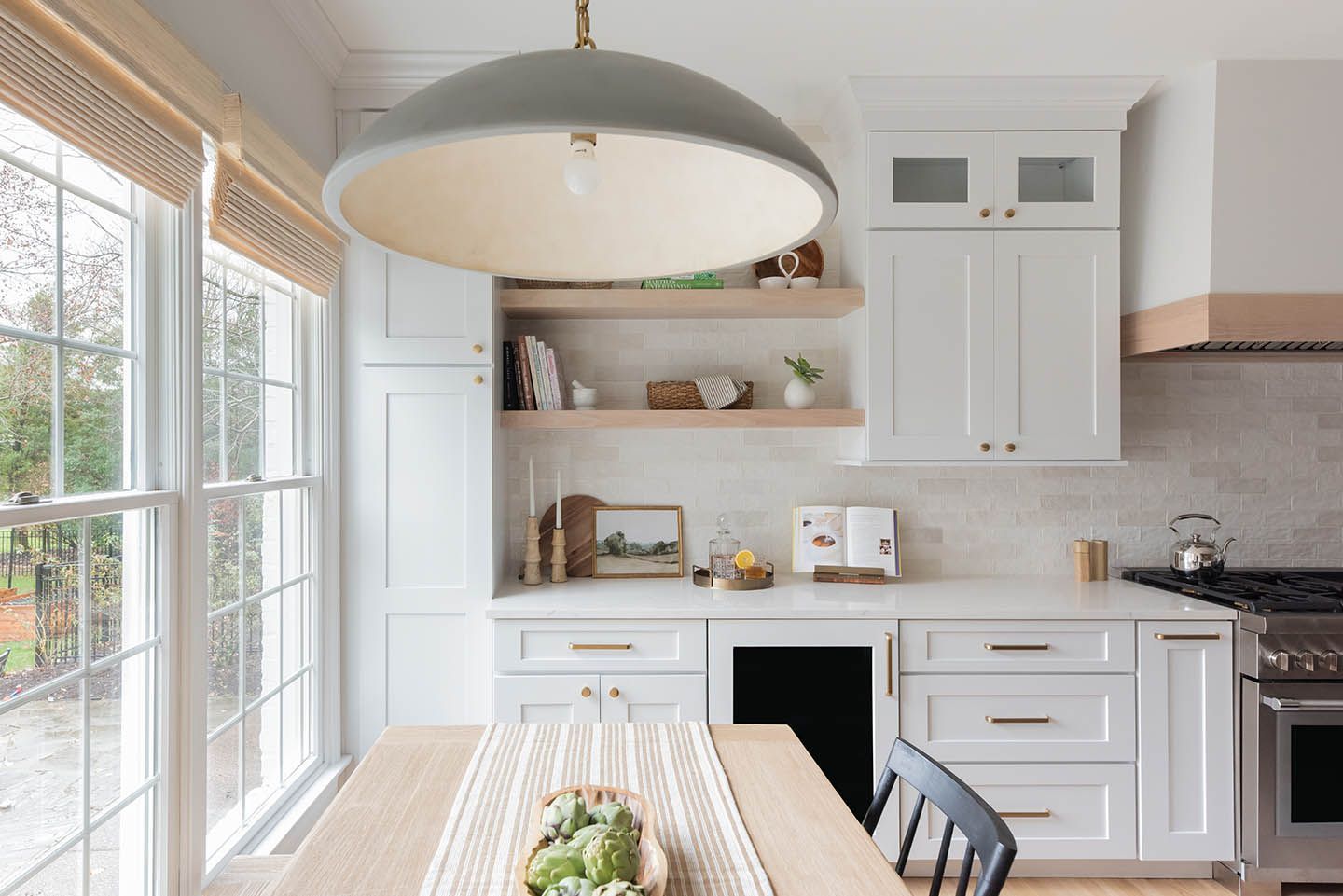 Bright kitchen with white cabinets, light wood shelves, and large window. A gray pendant light hangs overhead.