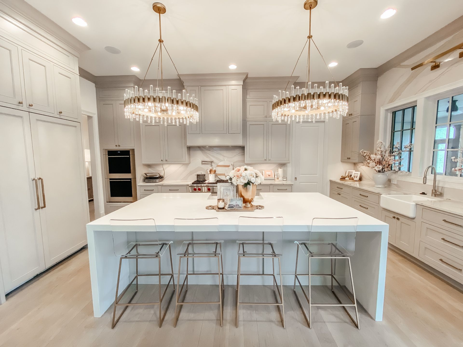 Modern kitchen with gray cabinets, white countertops, island with gold bar stools, and two crystal chandeliers.