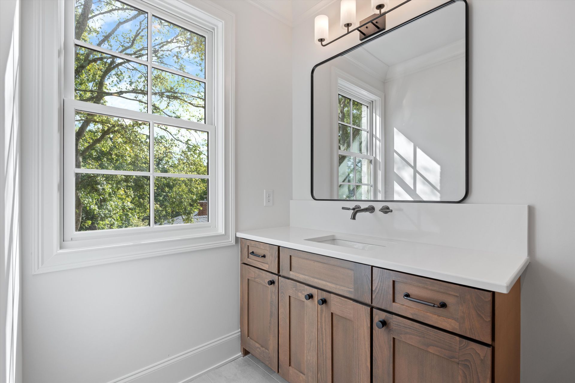 Bathroom with a wooden vanity, white countertop, large mirror, and window with a view of trees.