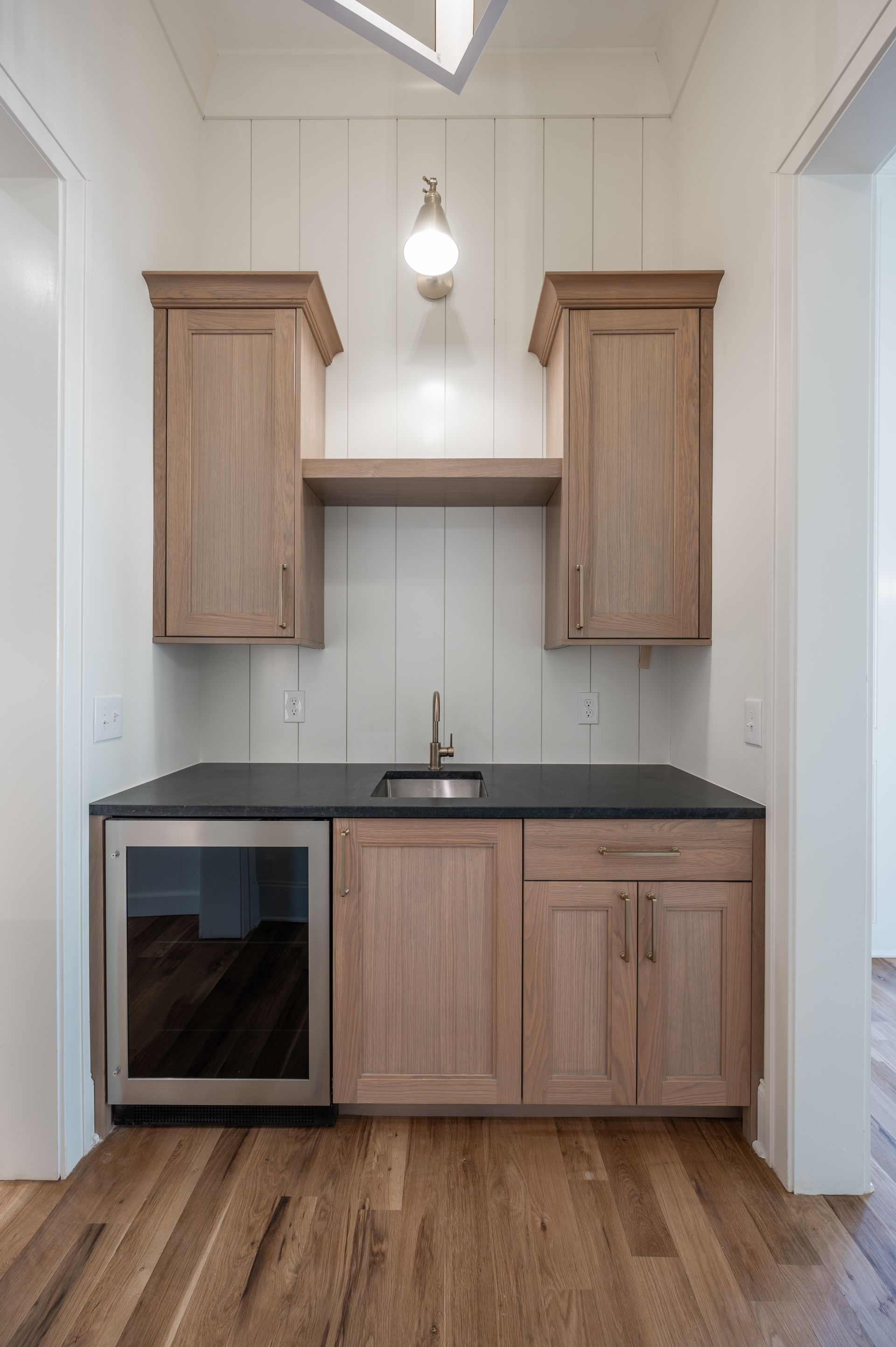 Built-in home bar with wood cabinets, black countertop, sink, and mini fridge. Wooden floor.