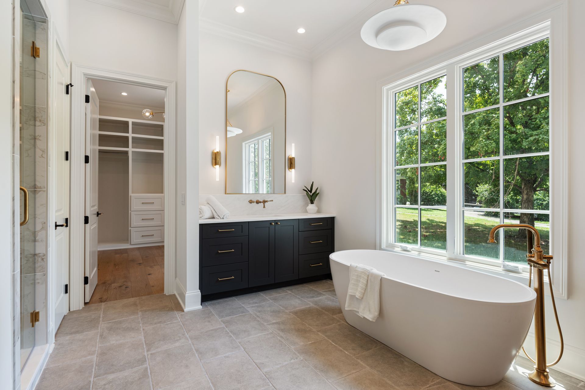 Modern bathroom with white walls, black vanity, gold accents, and large window overlooking greenery.