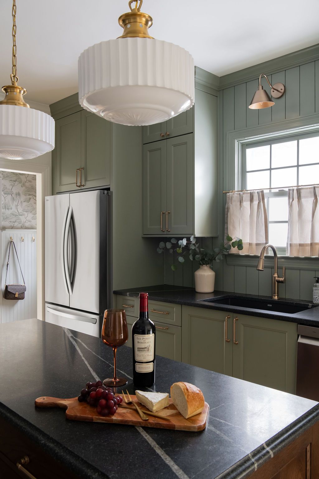 Kitchen with sage green cabinets, black countertop island, wine, cheese, and pendant lights.