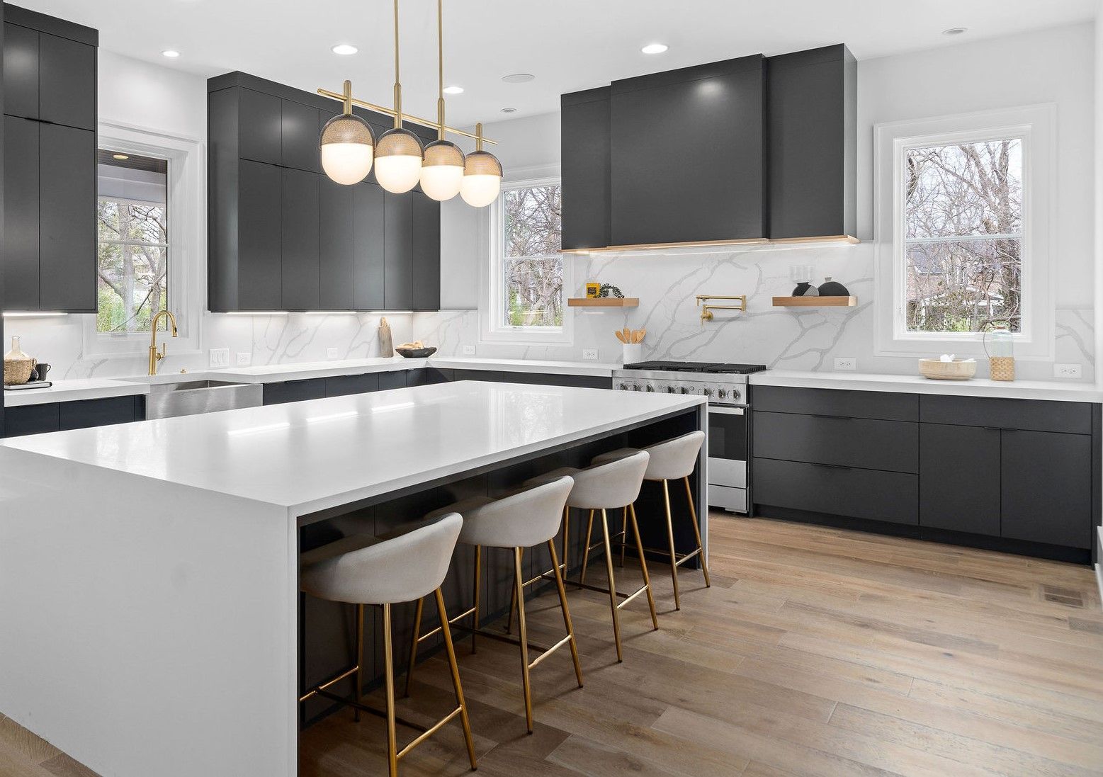 Modern kitchen with dark gray cabinets, white countertops, island with gold-legged stools, and wooden floor.