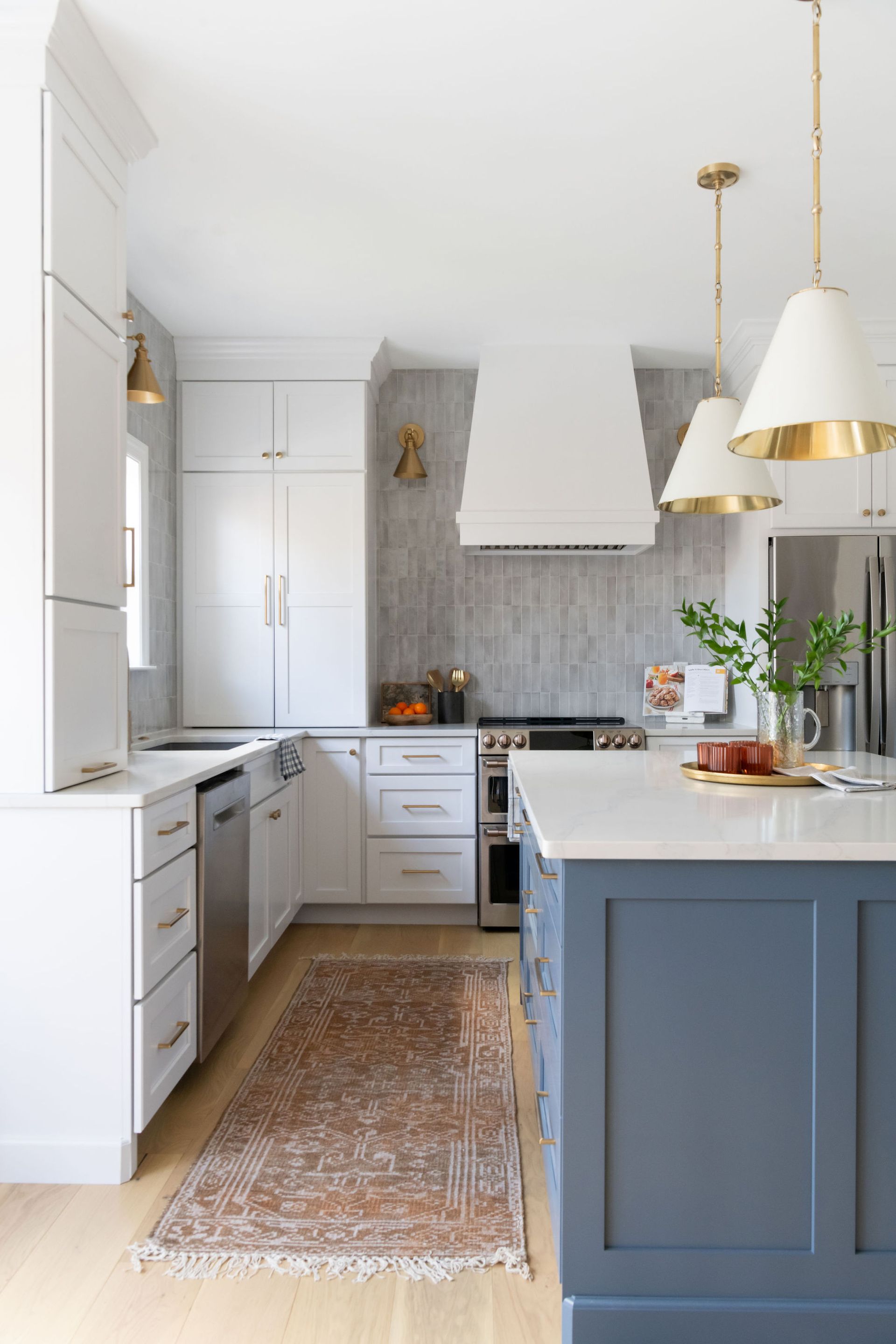 White kitchen with blue island, patterned rug, gold pendant lights, and stainless steel appliances.