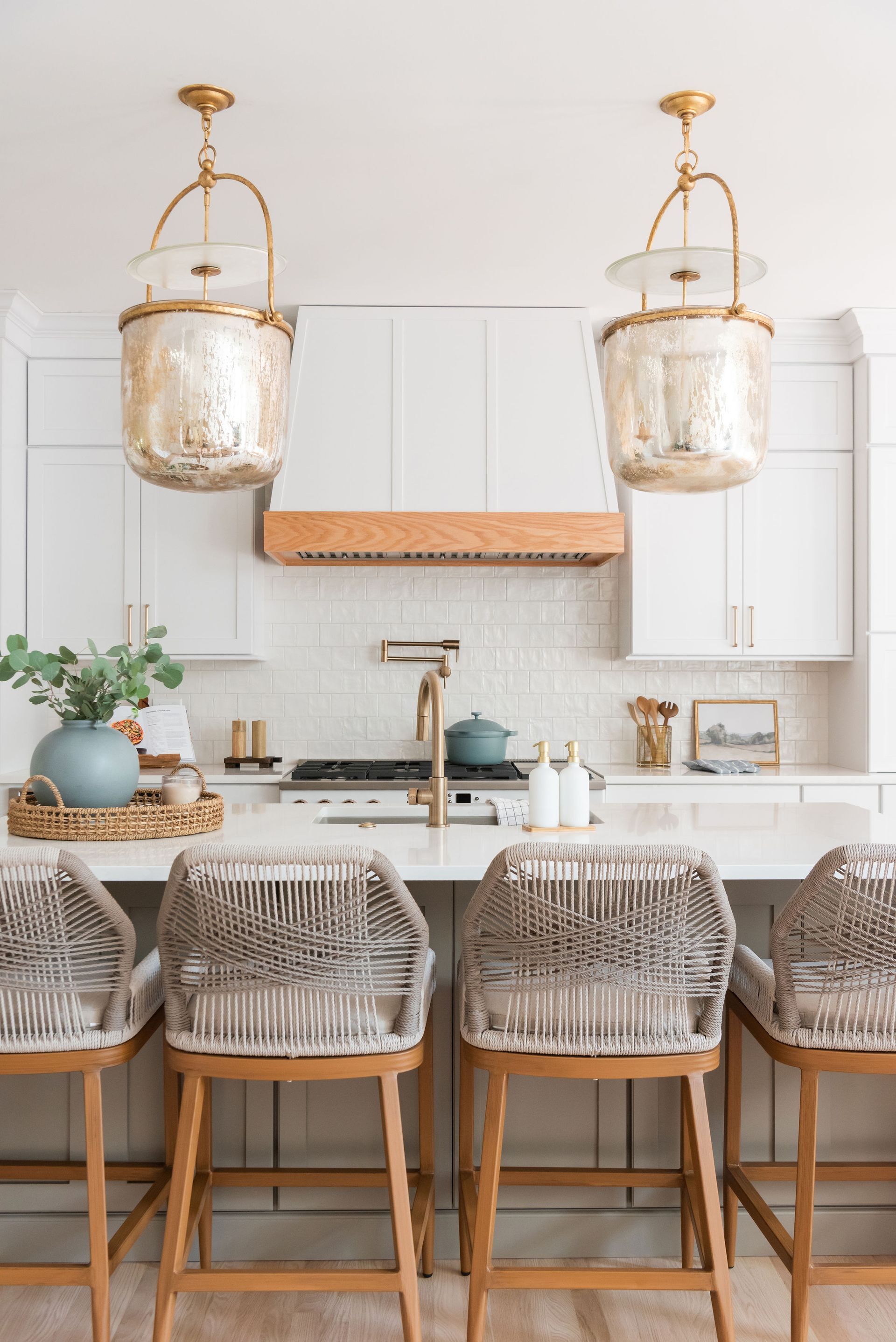 Kitchen with white cabinets, gold light fixtures, wooden range hood, and woven bar stools.