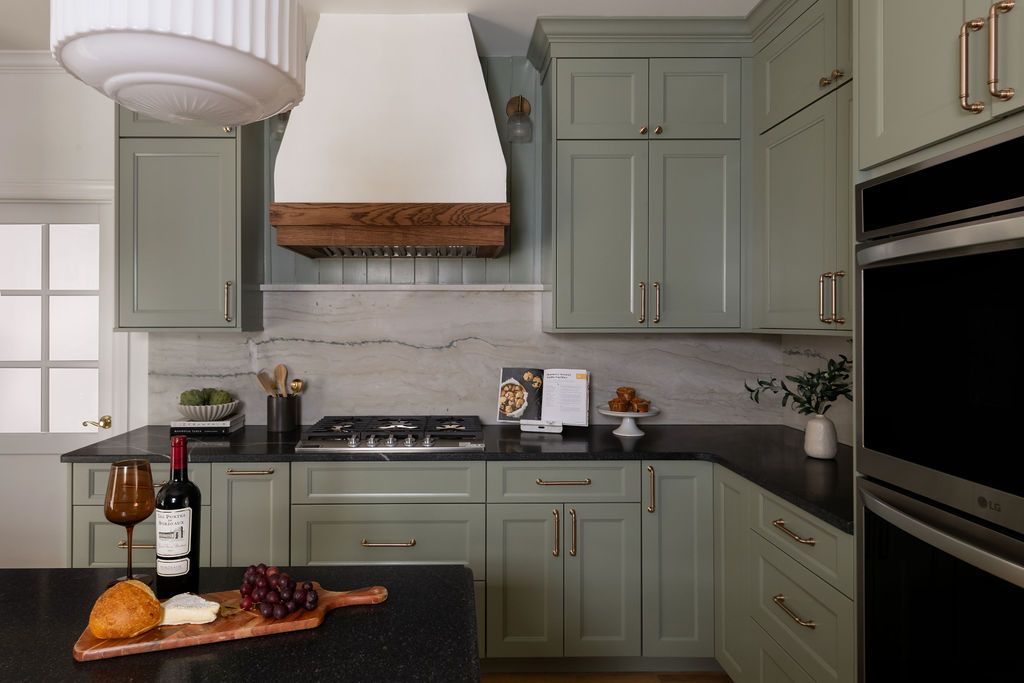 Kitchen with sage green cabinets, black countertops, and a rustic range hood. Wine and cheese on island.