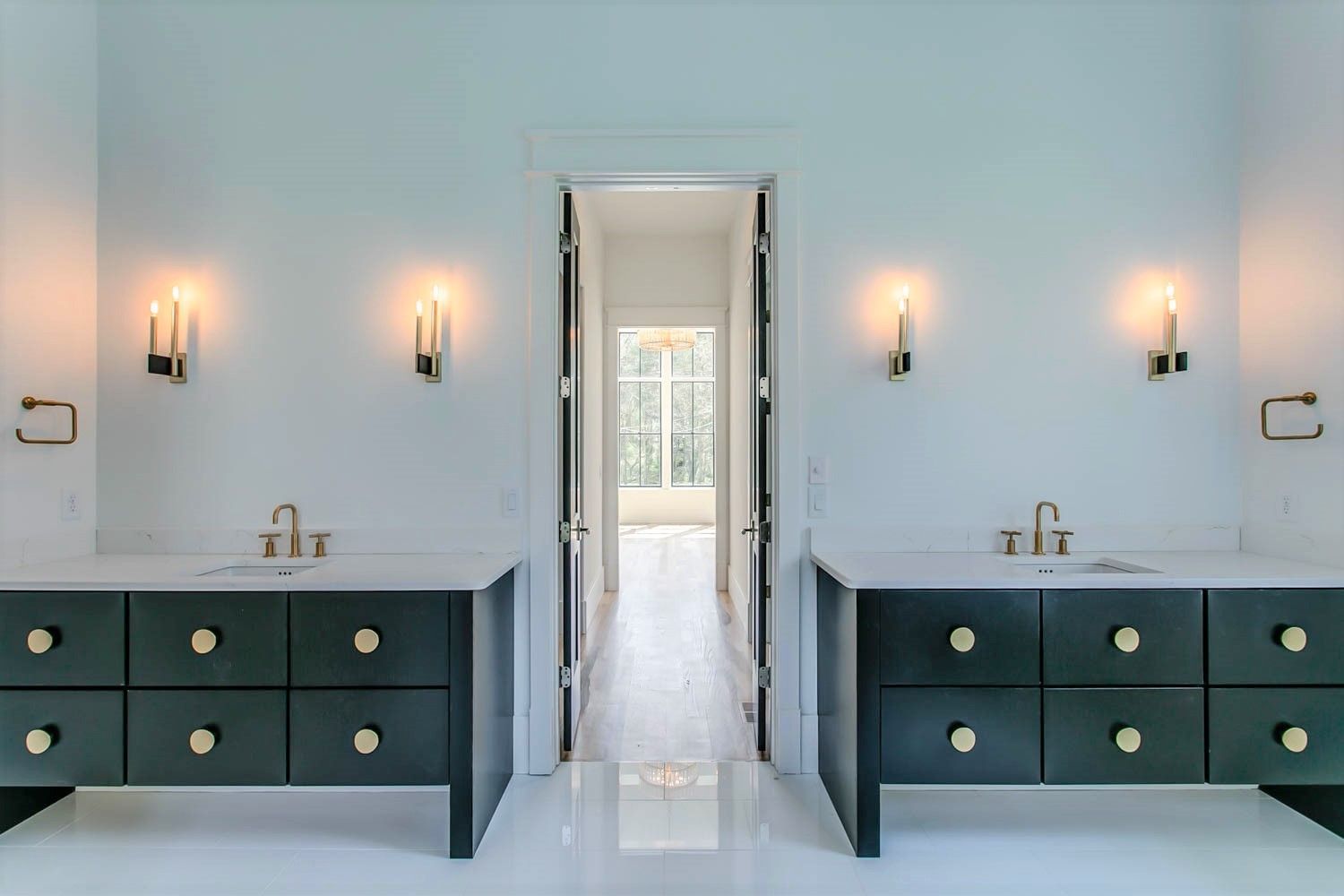 Modern bathroom with two dark vanities, gold fixtures, sconces, and a doorway with natural light.