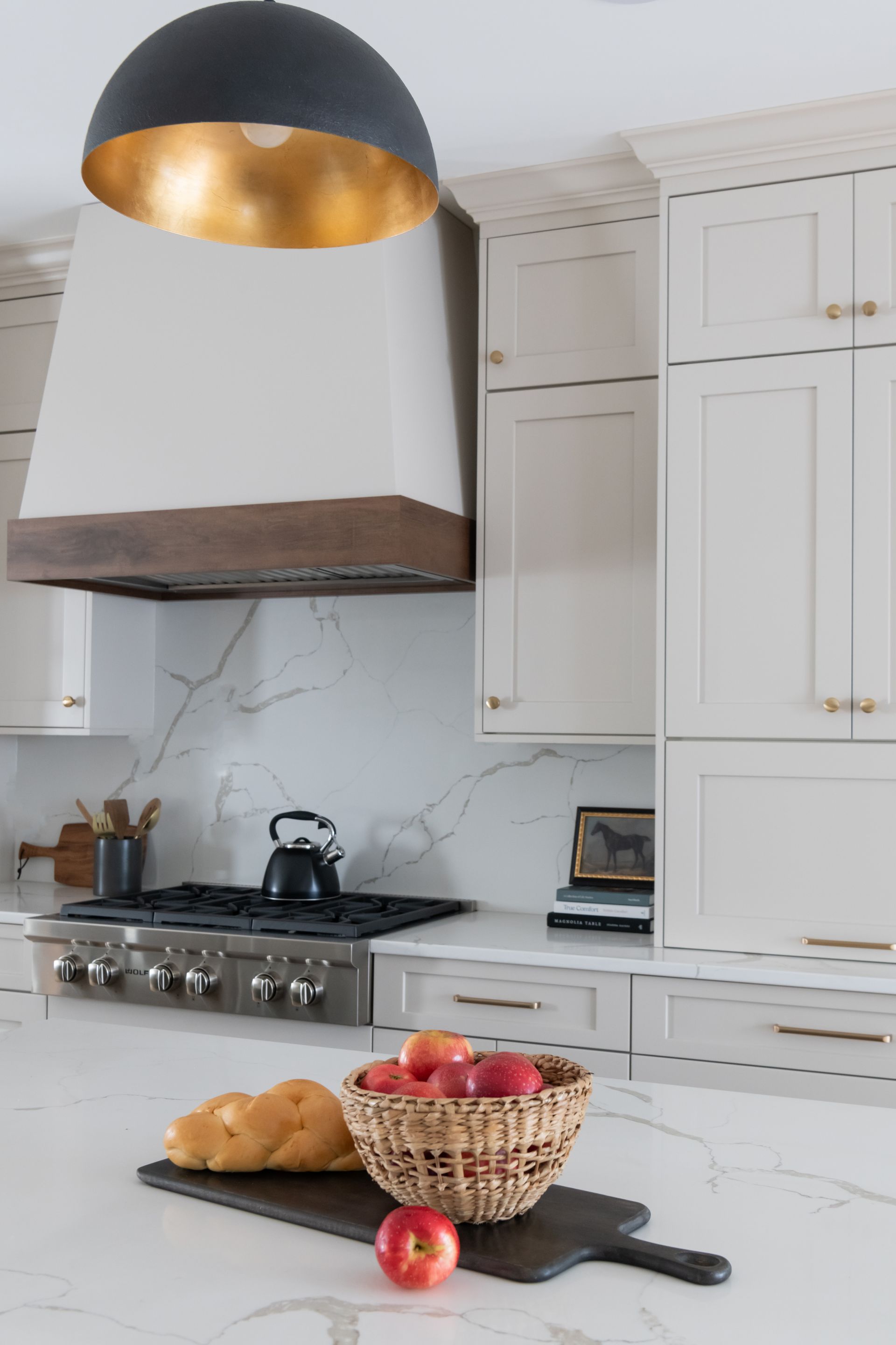 Kitchen with light cabinets, marble countertops, and a black and gold pendant light. A bowl of apples sits on a cutting board.
