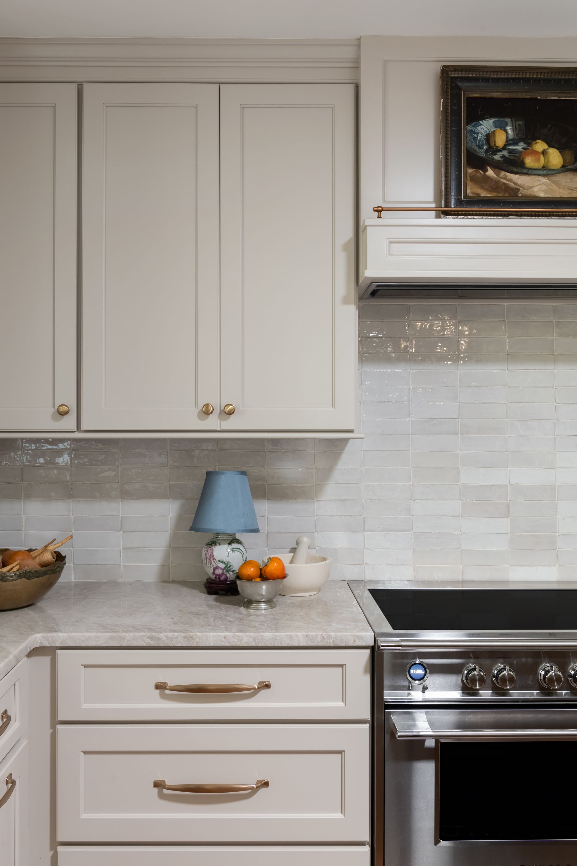Kitchen with light-colored cabinets, white tile backsplash, a stove, and countertop decor including a lamp.