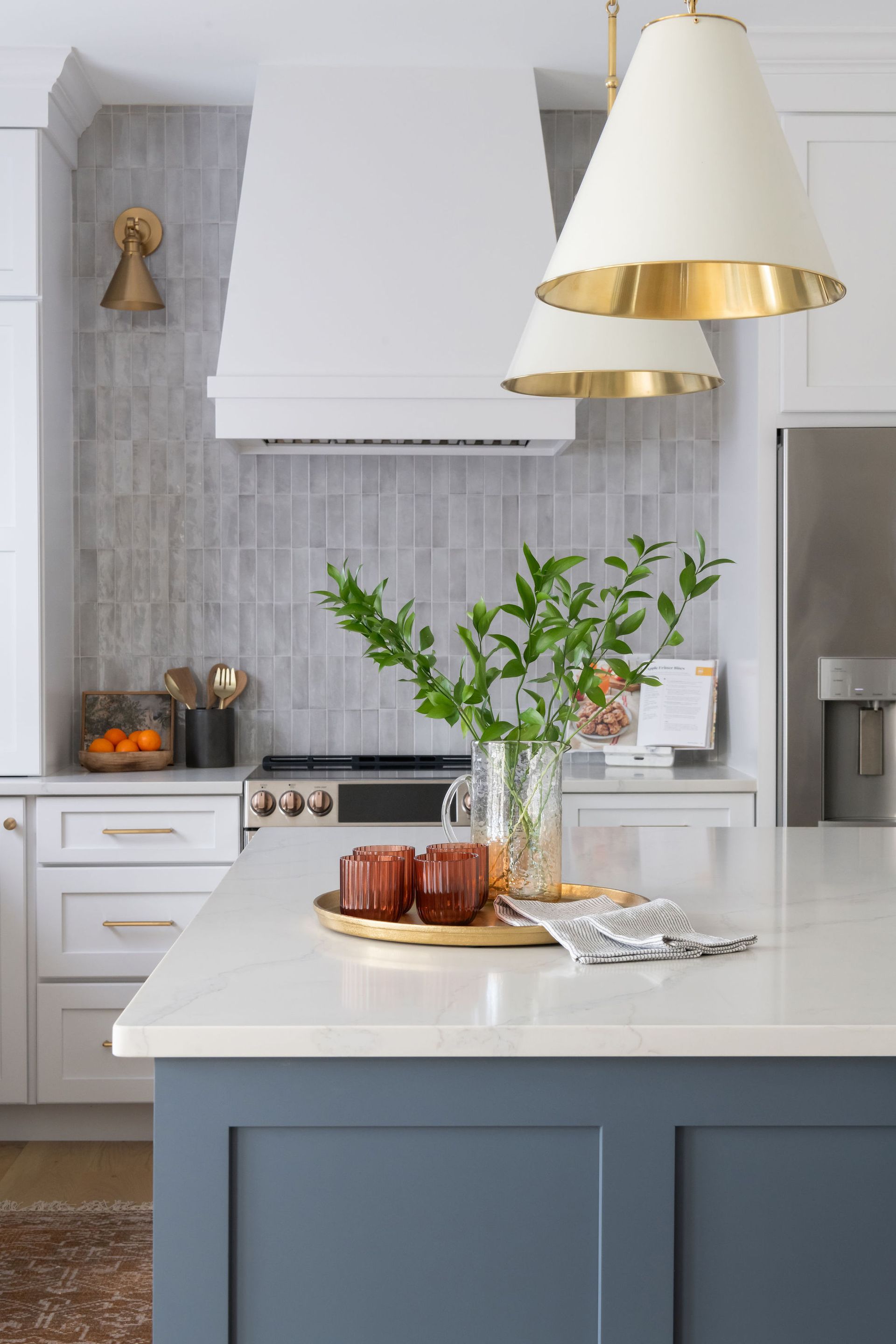 White and blue kitchen with island. Overhead lights, gray backsplash, greenery, and countertop decor.