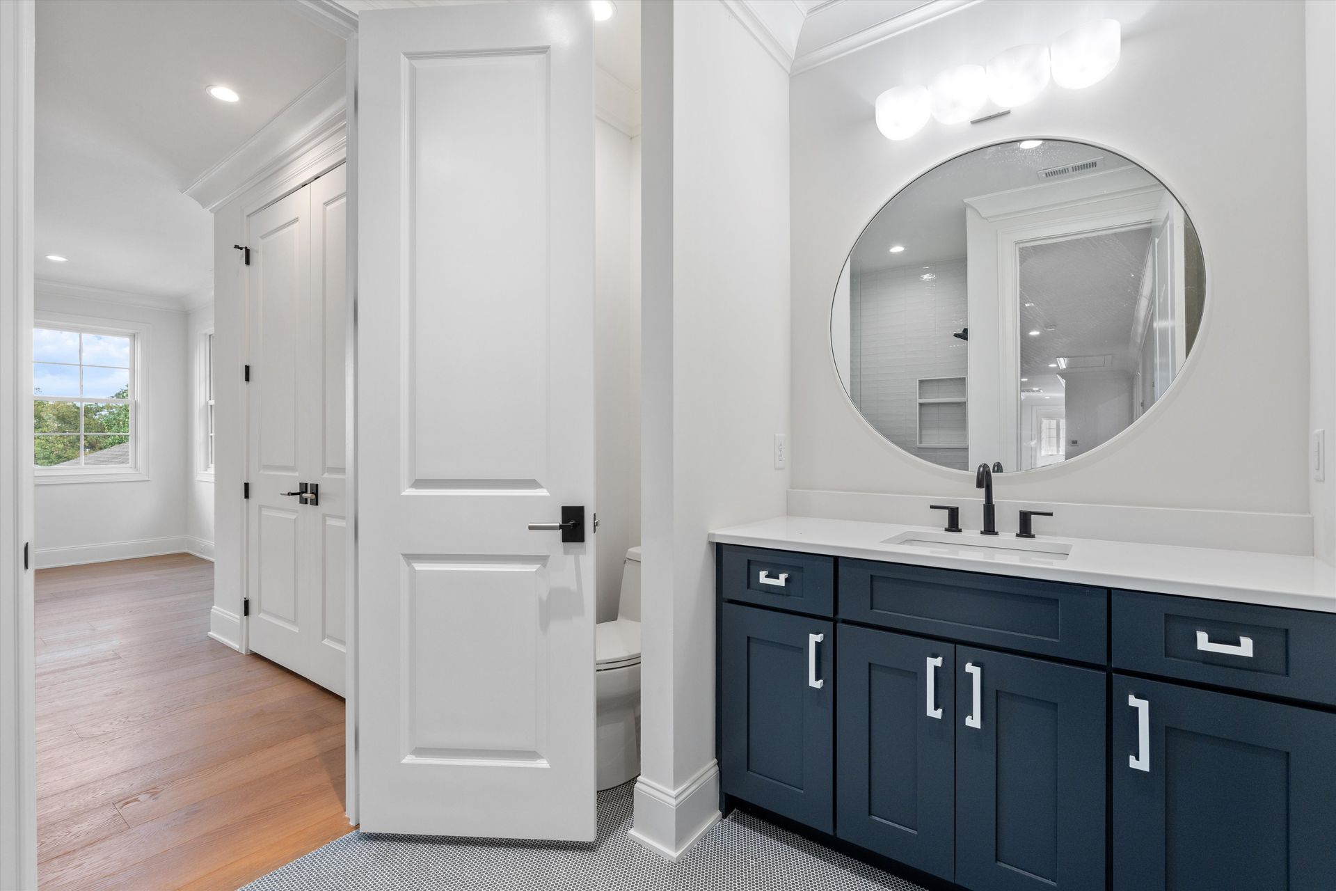 A bathroom with navy vanity, large round mirror, white walls, and a hallway with a door.