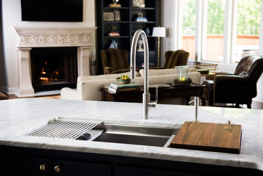 Kitchen sink with faucet, cutting board, and rack on a marble countertop; living room in the background.