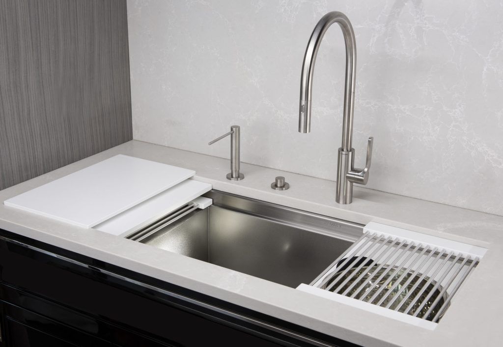 Stainless steel kitchen sink with faucet, cutting board, and dish rack on a white countertop.