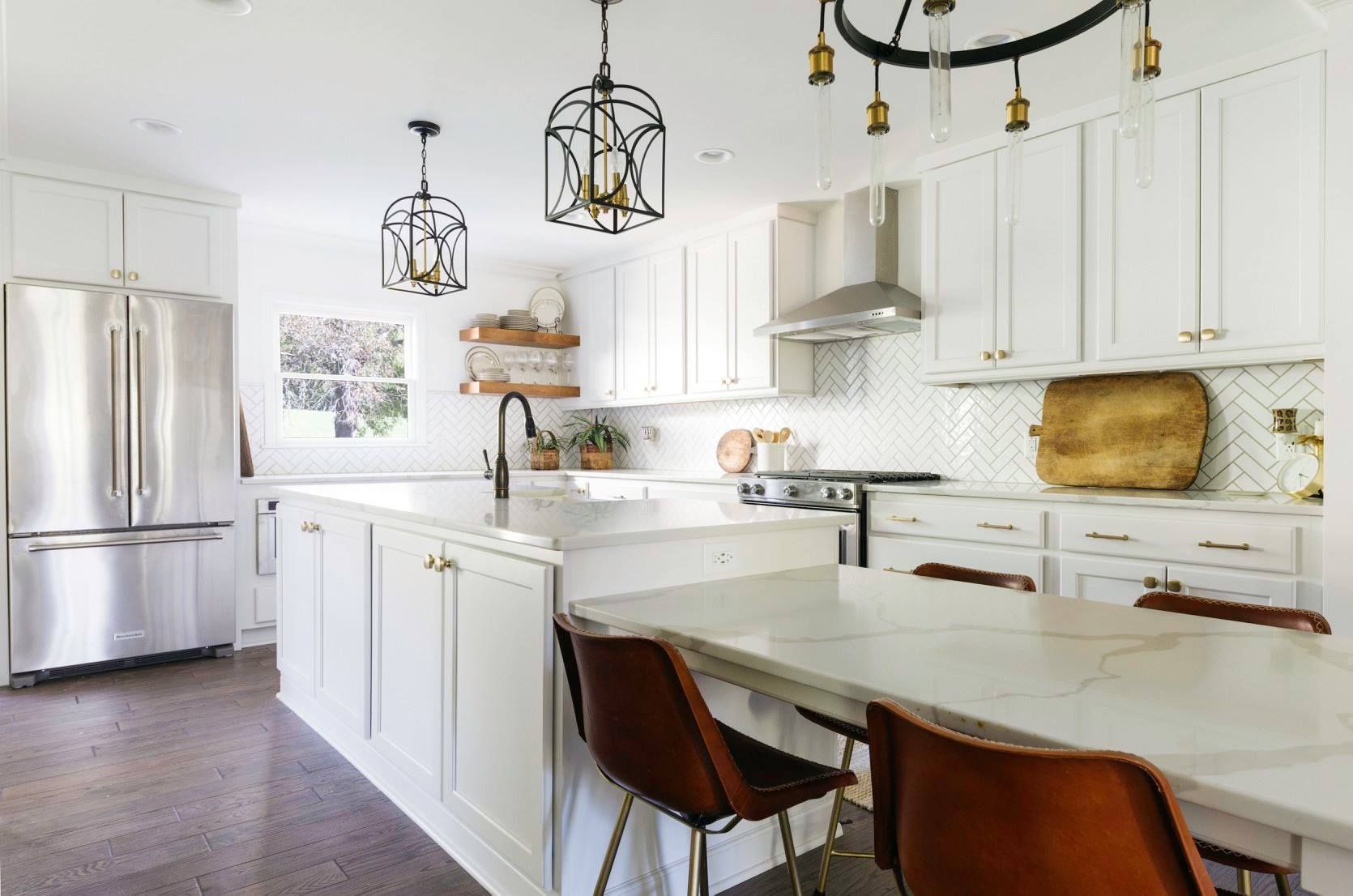 A modern white kitchen with a large island, stainless steel refrigerator, leather chairs, and hanging pendant lights.