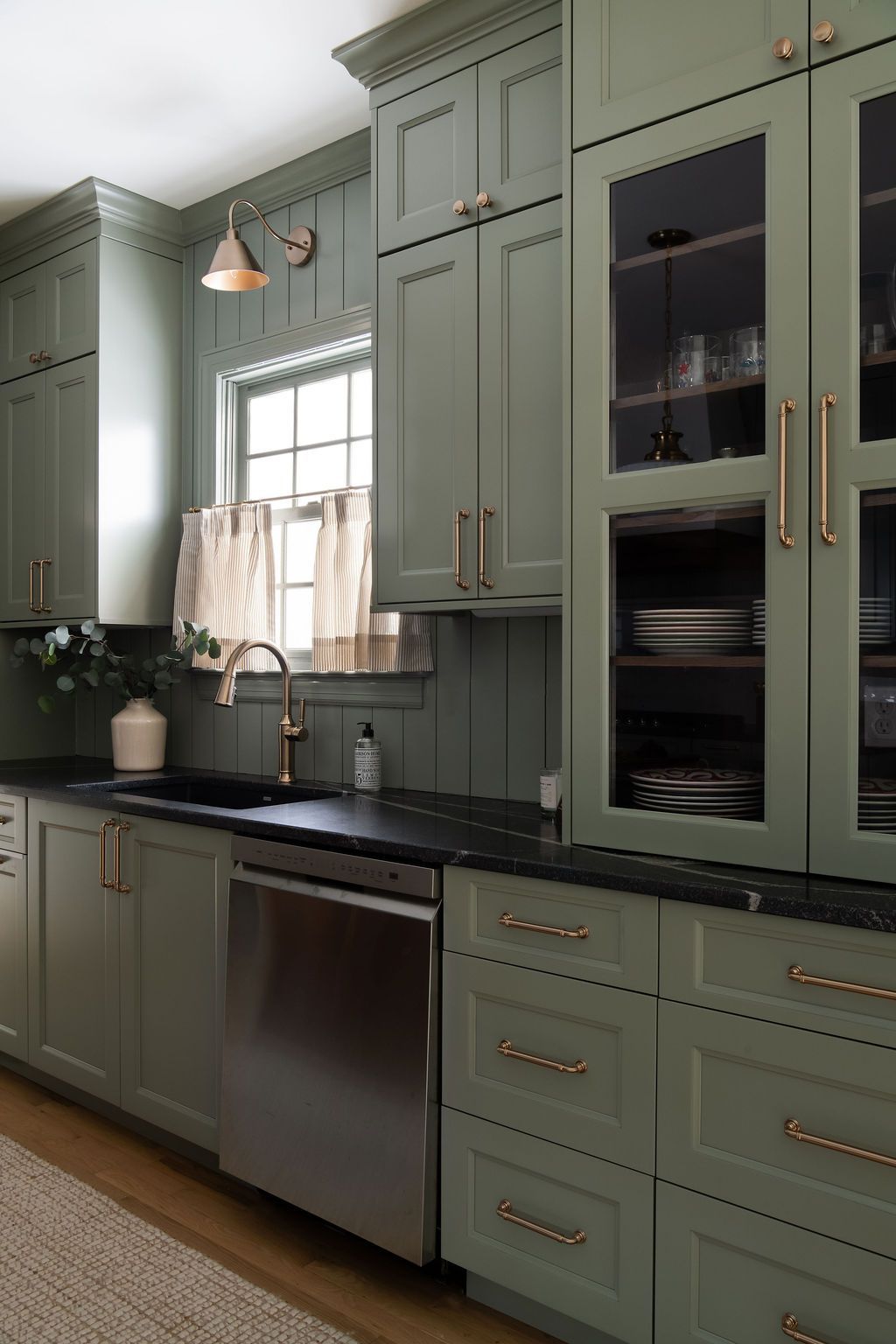 Sage green kitchen cabinets with brass hardware, a stainless steel dishwasher, black countertops, and a window above a sink.