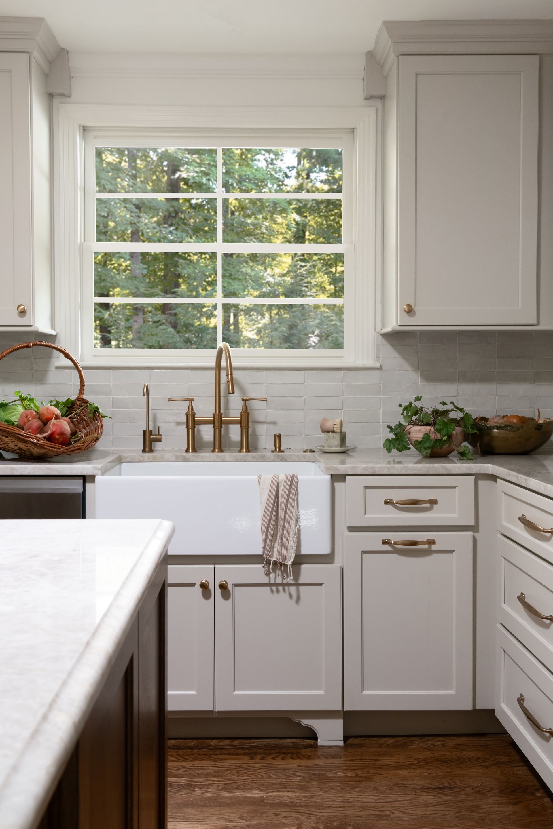 A bright kitchen with a white farmhouse sink, brass faucet, light grey cabinets, and a window overlooking green trees.