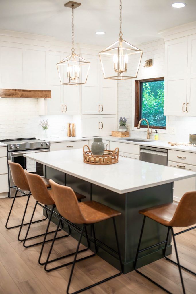 Modern white kitchen with a dark green island, gold pendant lights, and brown leather bar stools.