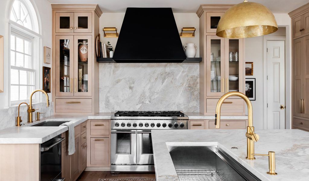 Elegant kitchen with light wood cabinets, marble backsplash, black range hood, and gold fixtures.