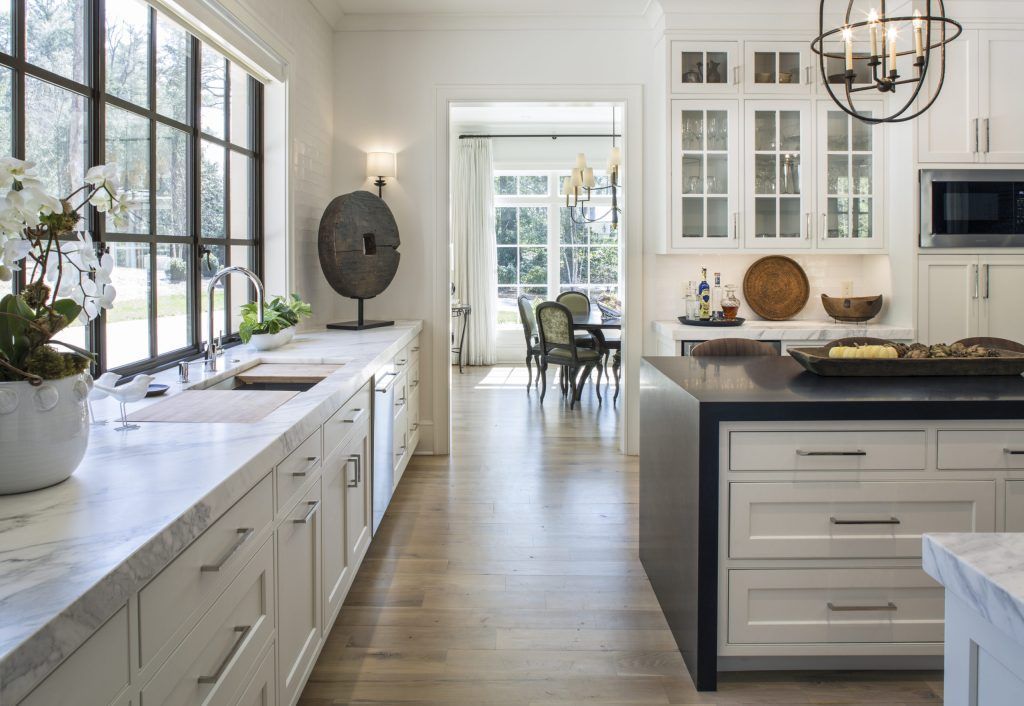 Modern white kitchen with marble countertops, large windows, and an island leading to a dining room.