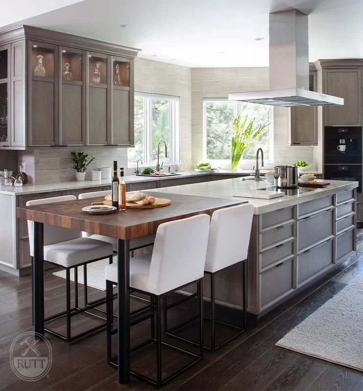Modern kitchen with gray cabinetry, large island, butcher block table, white bar stools, and stainless steel range hood.