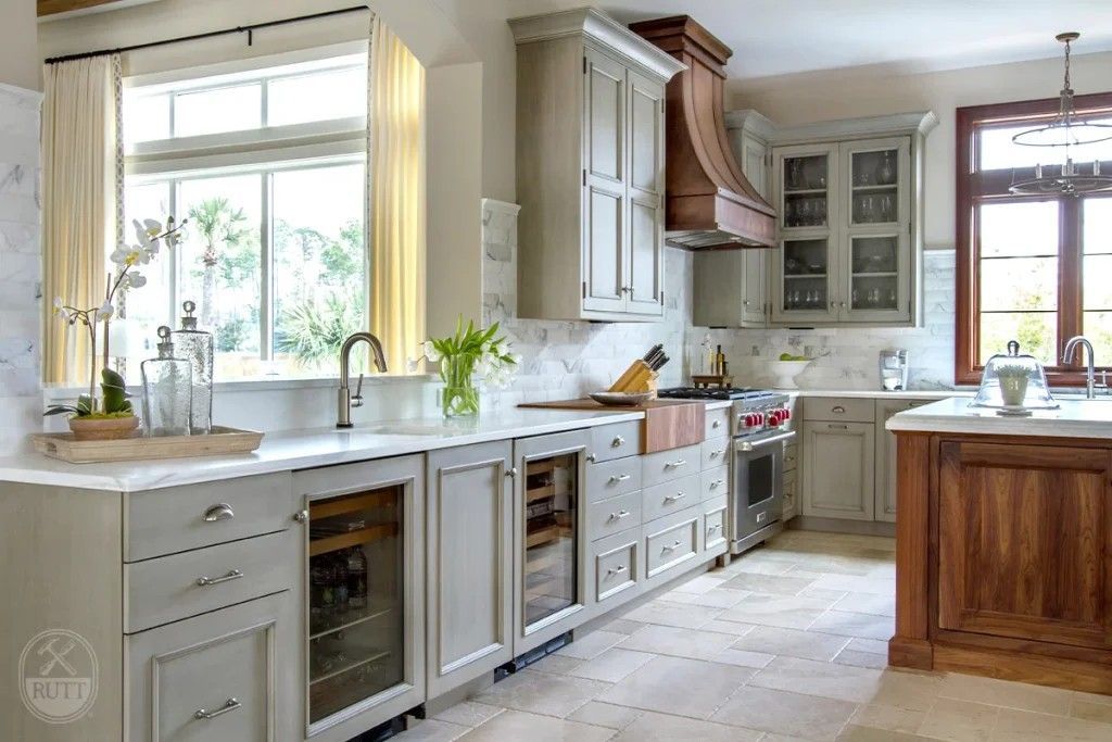 Elegant kitchen with gray cabinets, wooden range hood, marble countertops, and a large window.