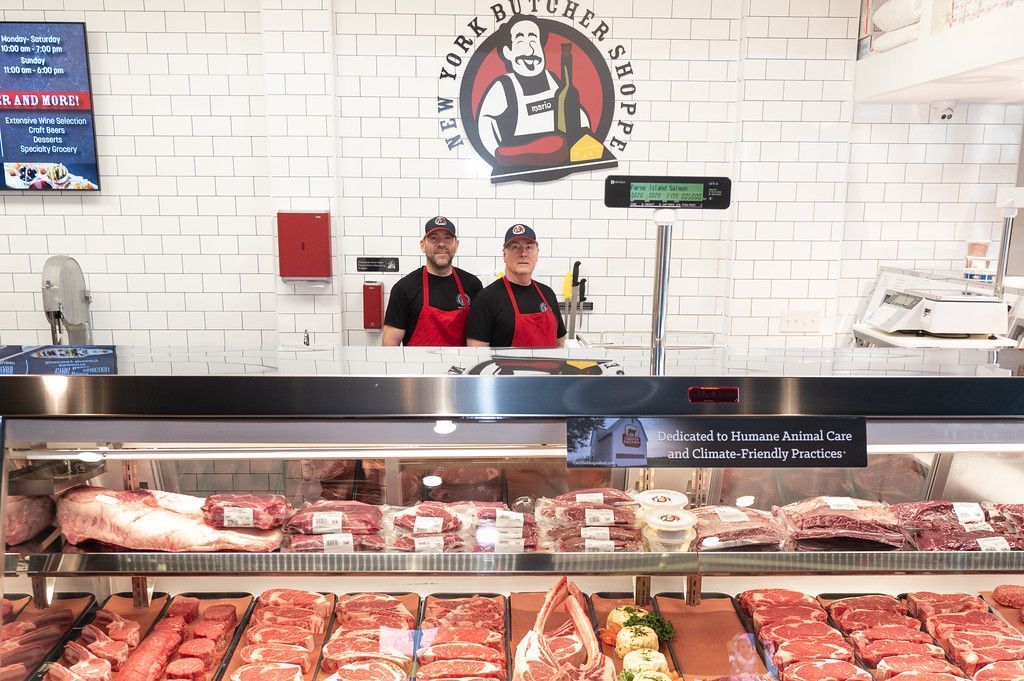 Two butchers smiling behind a glass display case full of various cuts of meat at the New York Butcher Shoppe.