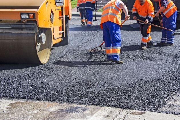 Road construction workers in high-visibility orange vests use tools to spread fresh asphalt near a yellow steamroller.