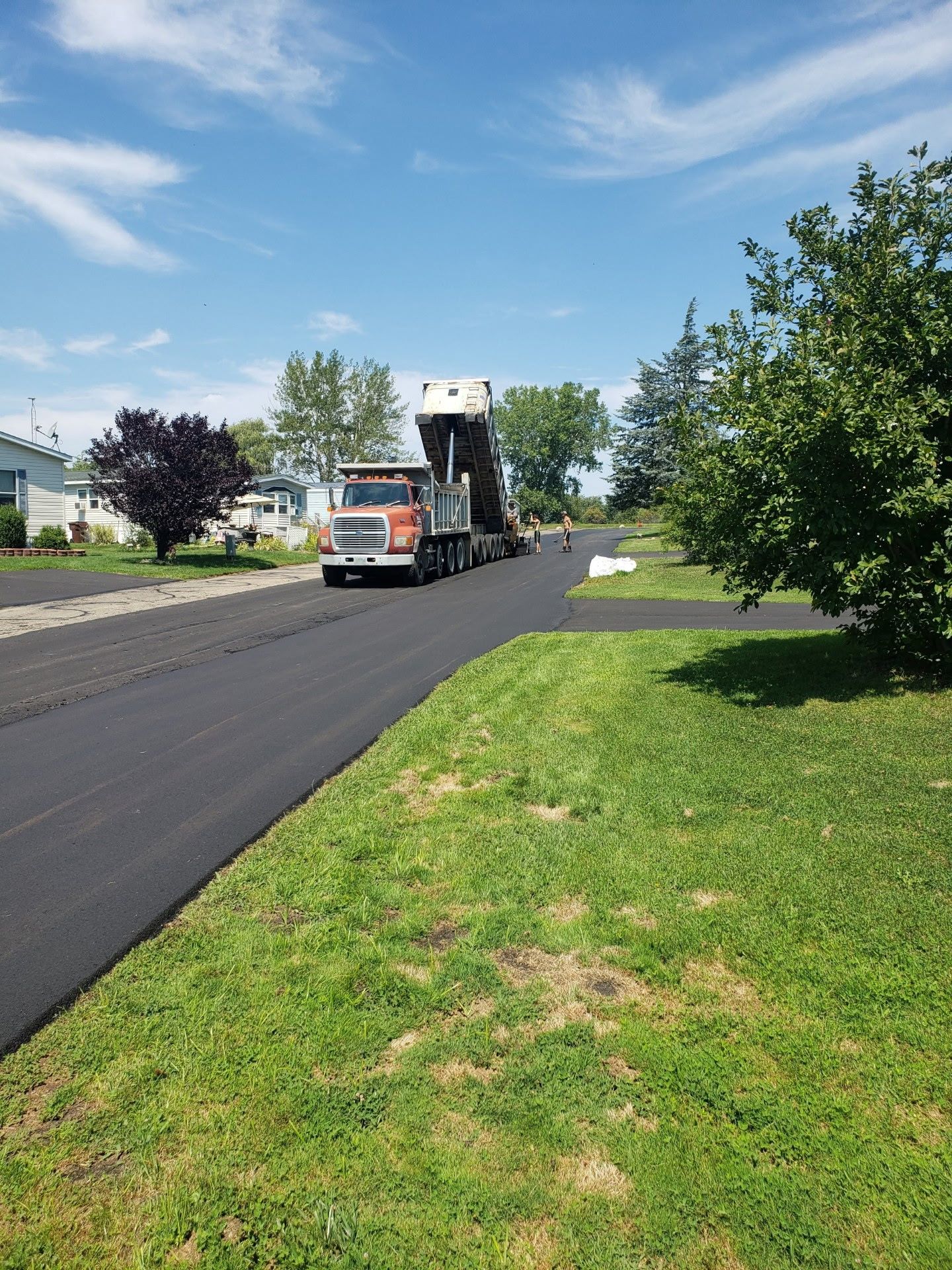 Truck carrying a large machine on a freshly paved road beside a grassy verge and trees under a blue sky
