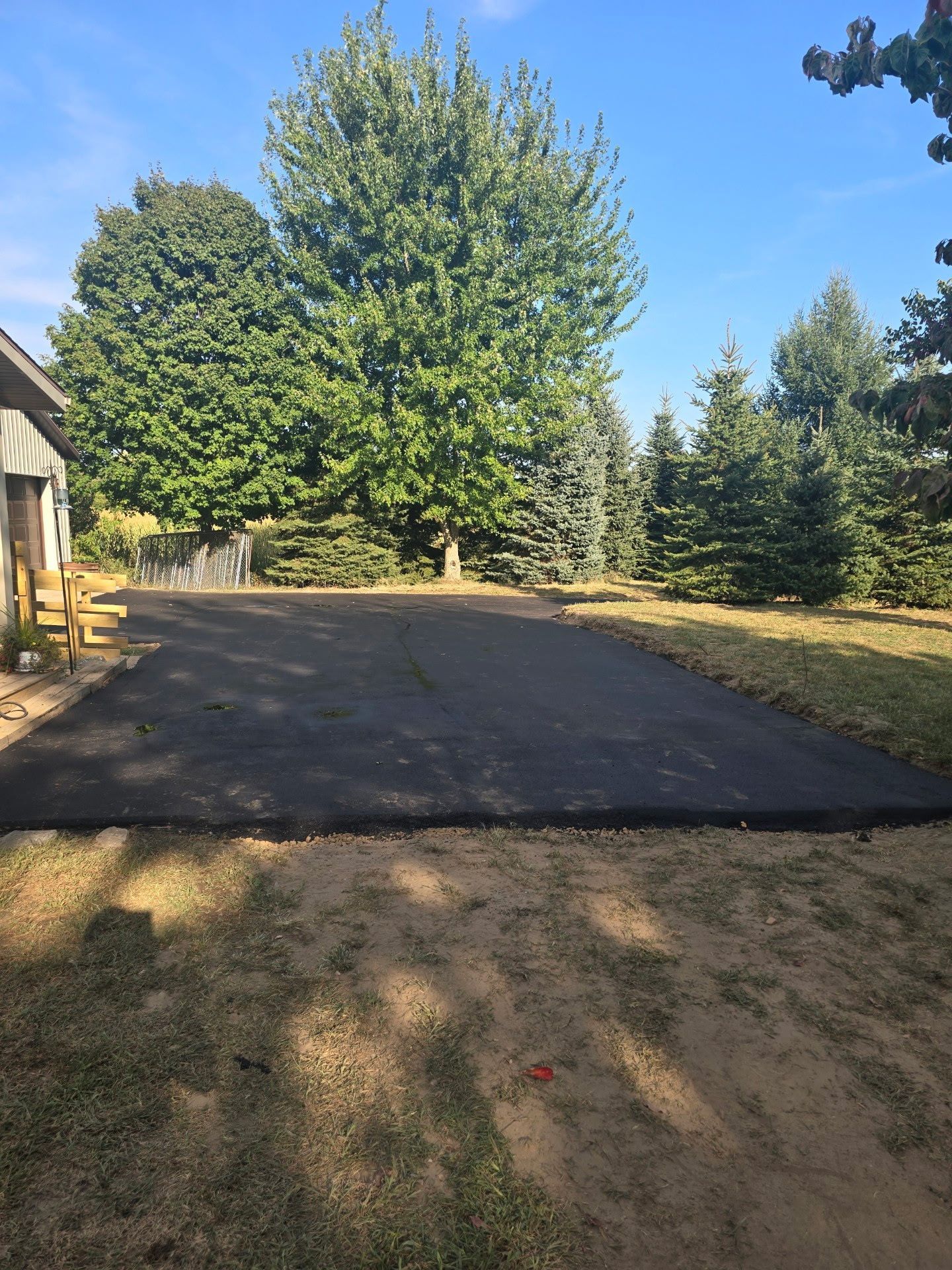 Driveway under construction beside a house, with fresh black asphalt and trees in the background.