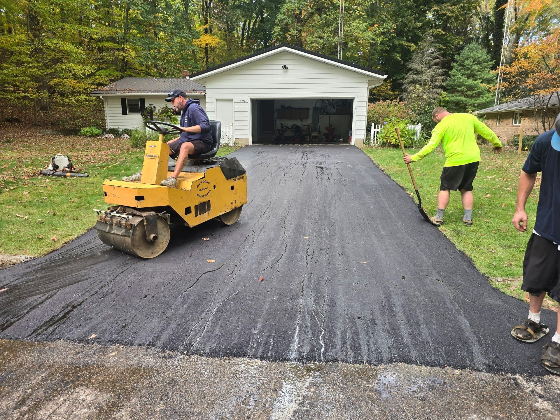Workers paving a driveway with a yellow roller in front of a garage, surrounded by trees
