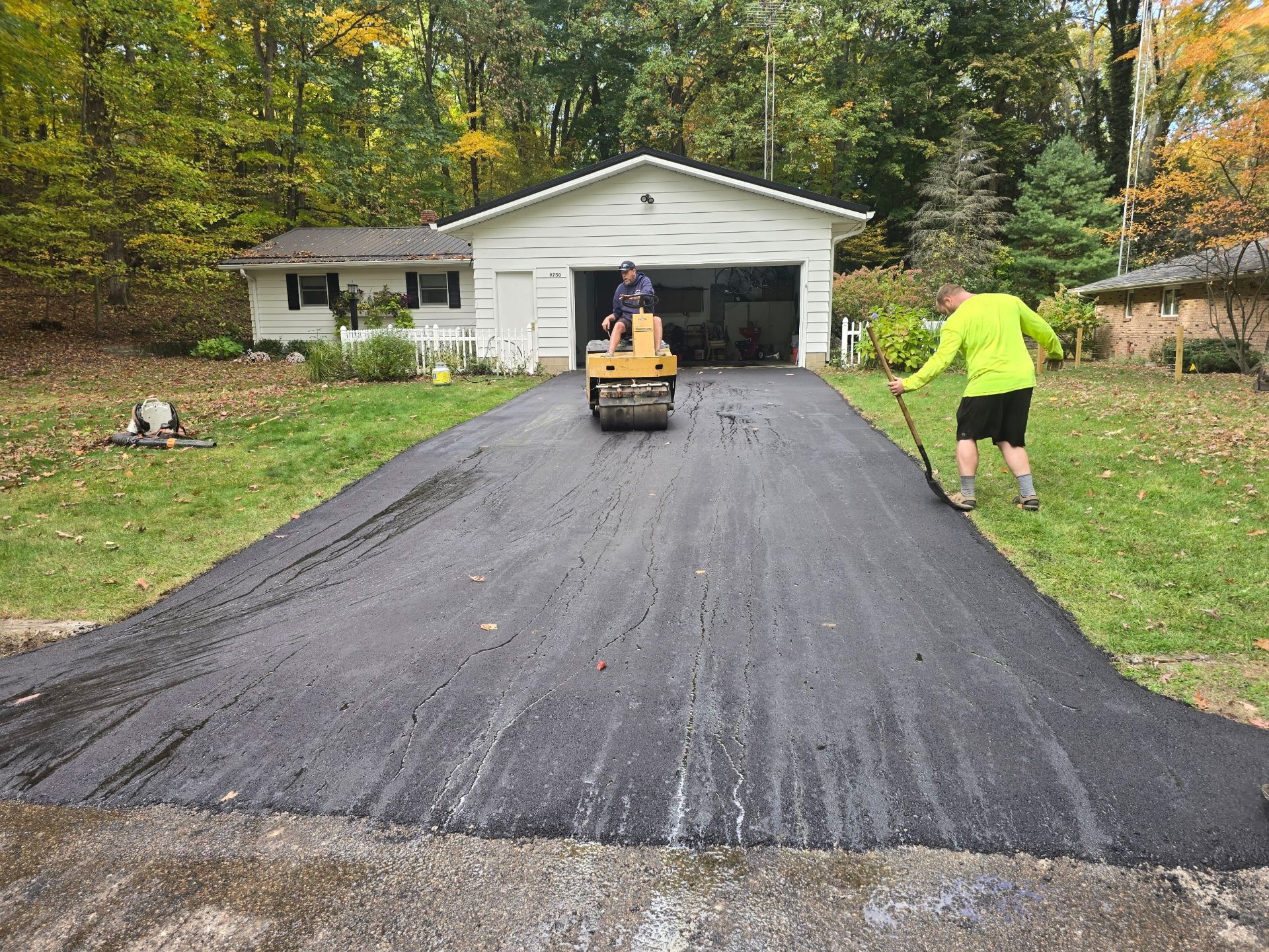 Two workers paving a long black driveway in front of a white garage, with a compact roller on the fresh asphalt.