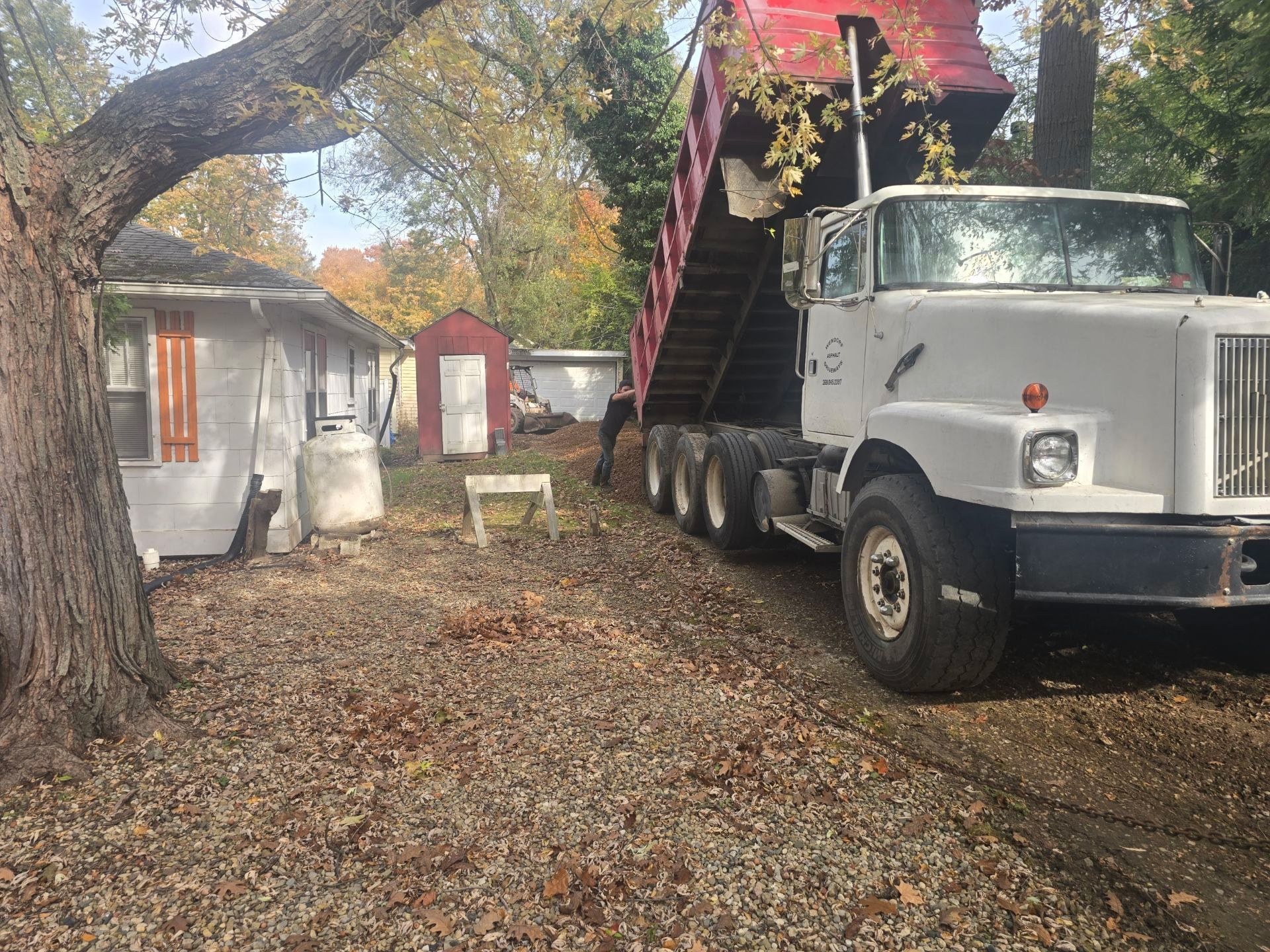 White dump truck with raised bed on a leaf-covered driveway beside a small house and trees.