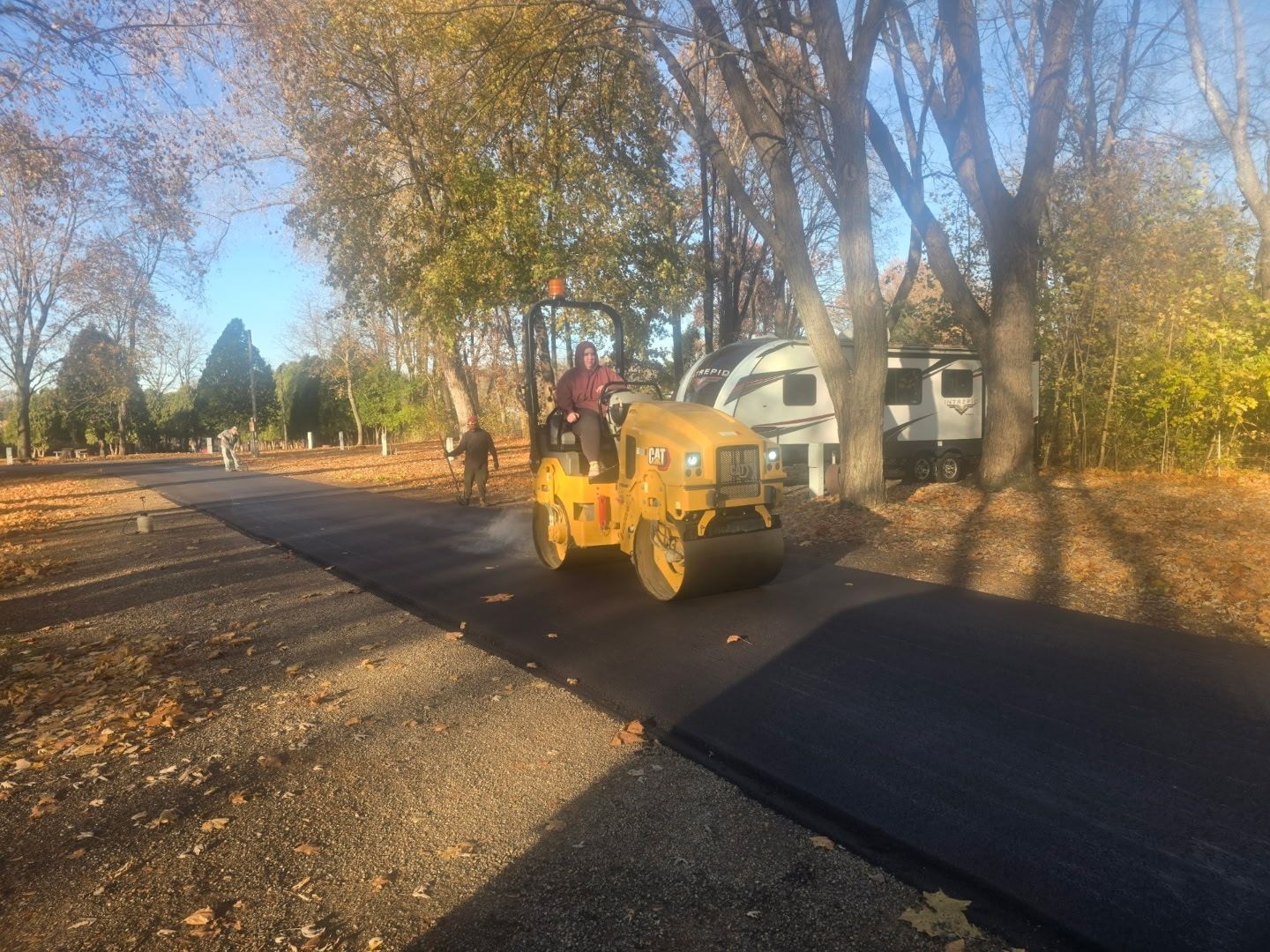 Yellow road roller paving a dark asphalt path beside trees and houses in fall