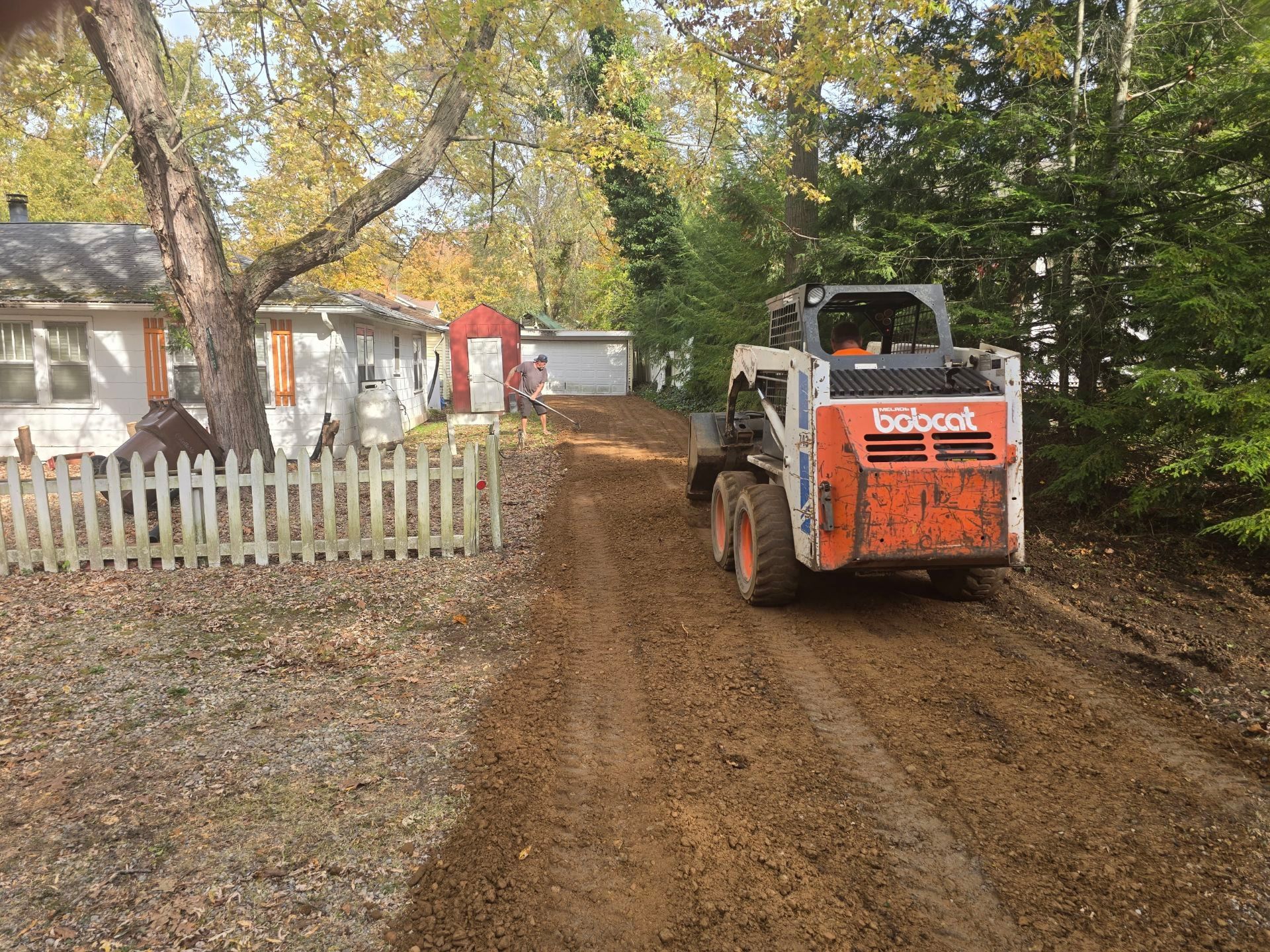 Orange skid steer on muddy driveway beside white picket fence and house in a leafy yard