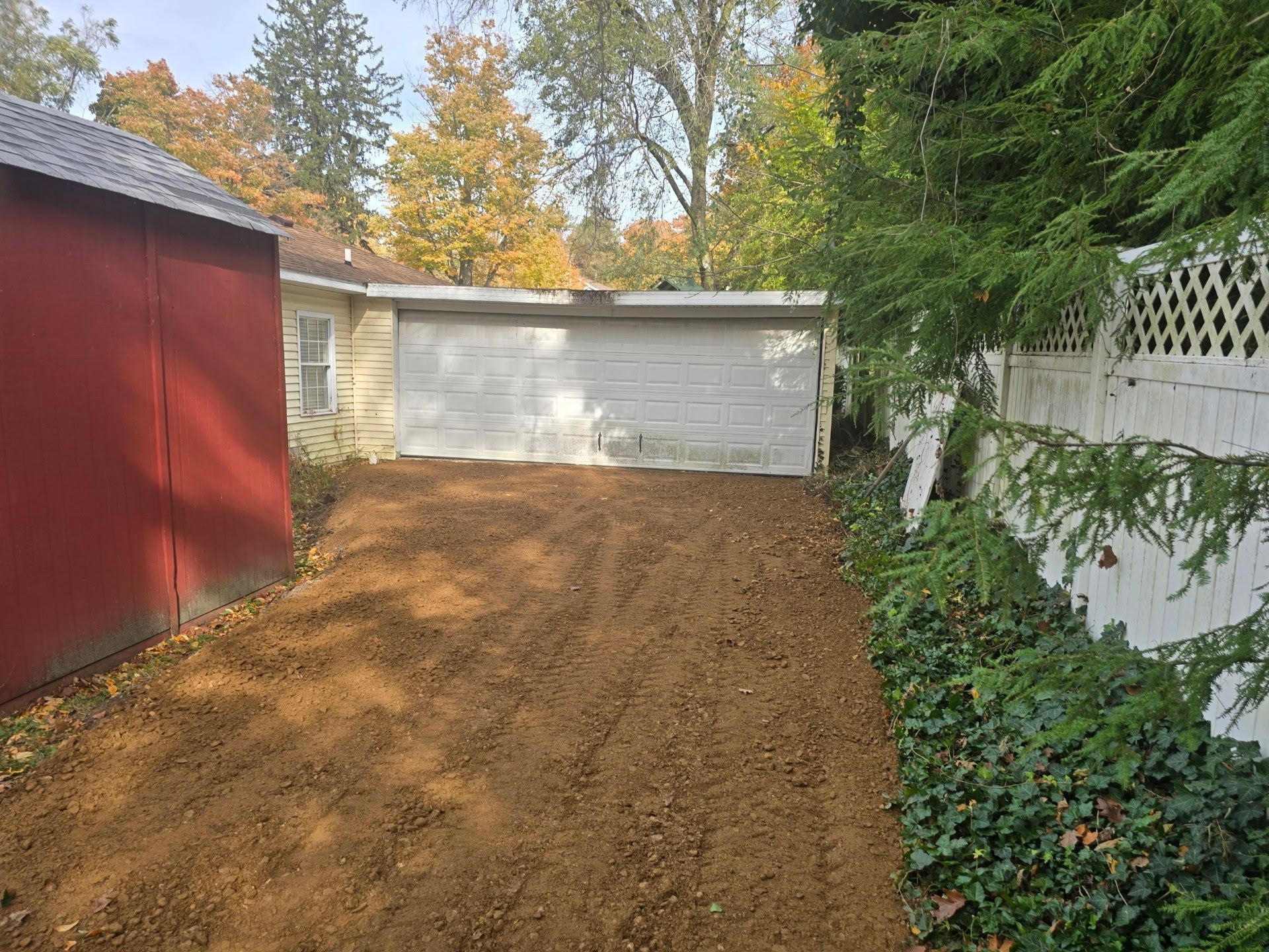Freshly graded dirt driveway leading to a white garage beside a red shed and white fence in a wooded yard