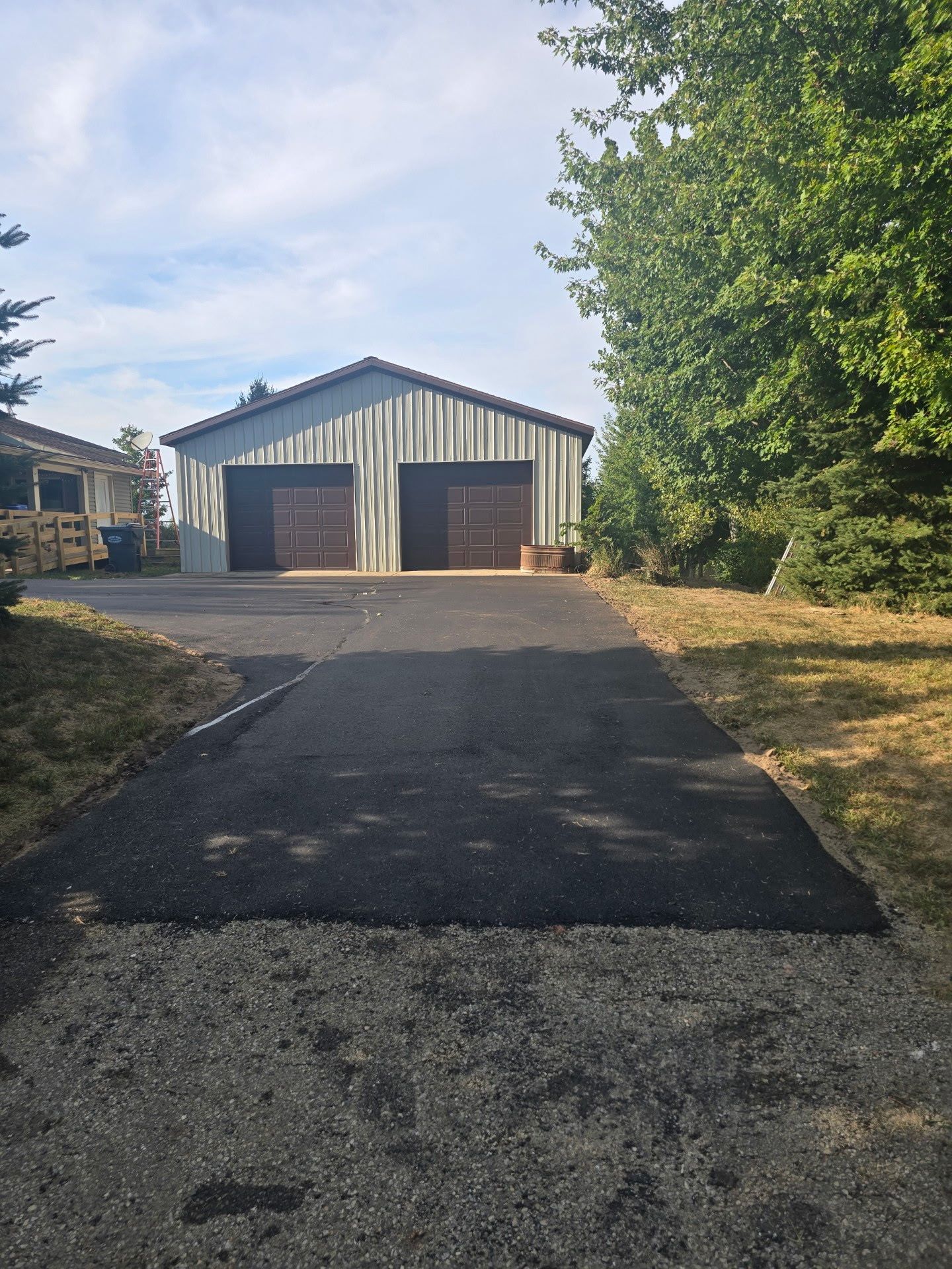 Driveway leading to a metal garage with two wooden doors, framed by trees and a blue sky.