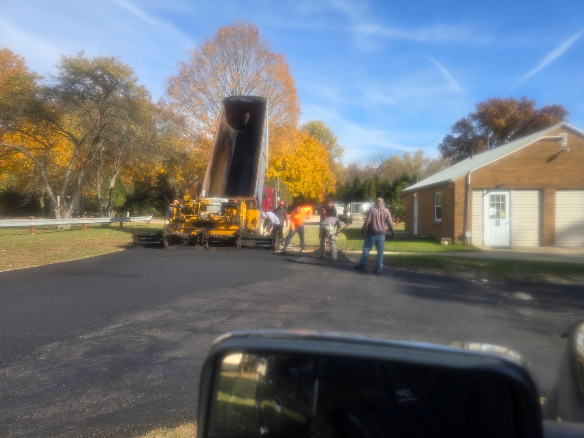 Workers and a raised dump truck on a paved road beside a brick building in autumn