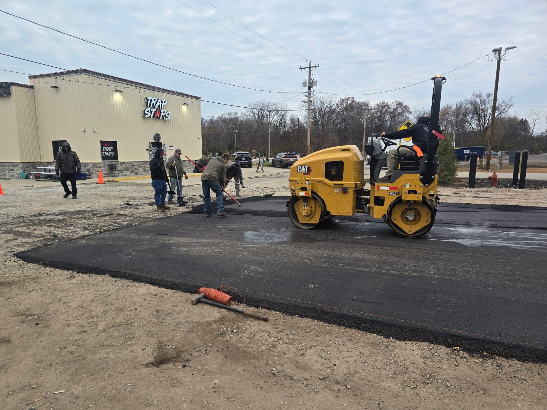 Workers paving a parking lot with a yellow roller near a beige building on a cloudy day