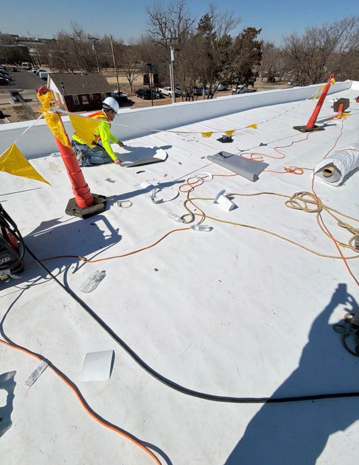 Roofer in safety gear on a white rooftop, near safety cones and ropes, under a sunny sky.