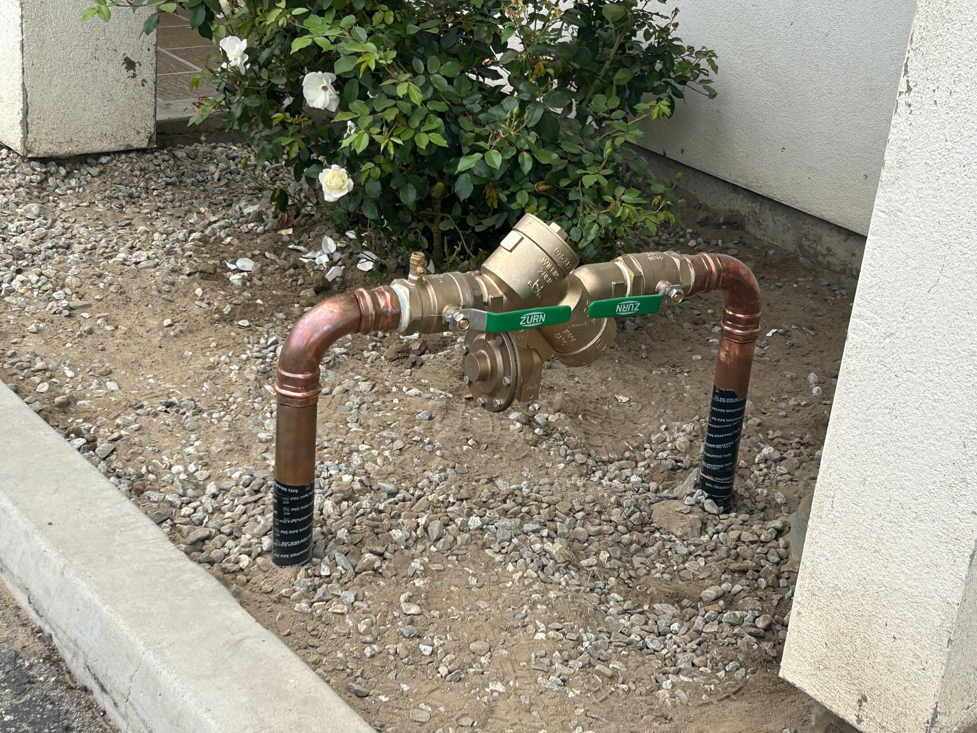 Copper irrigation pipes with green valves in a gravel bed beside a building corner and shrubs