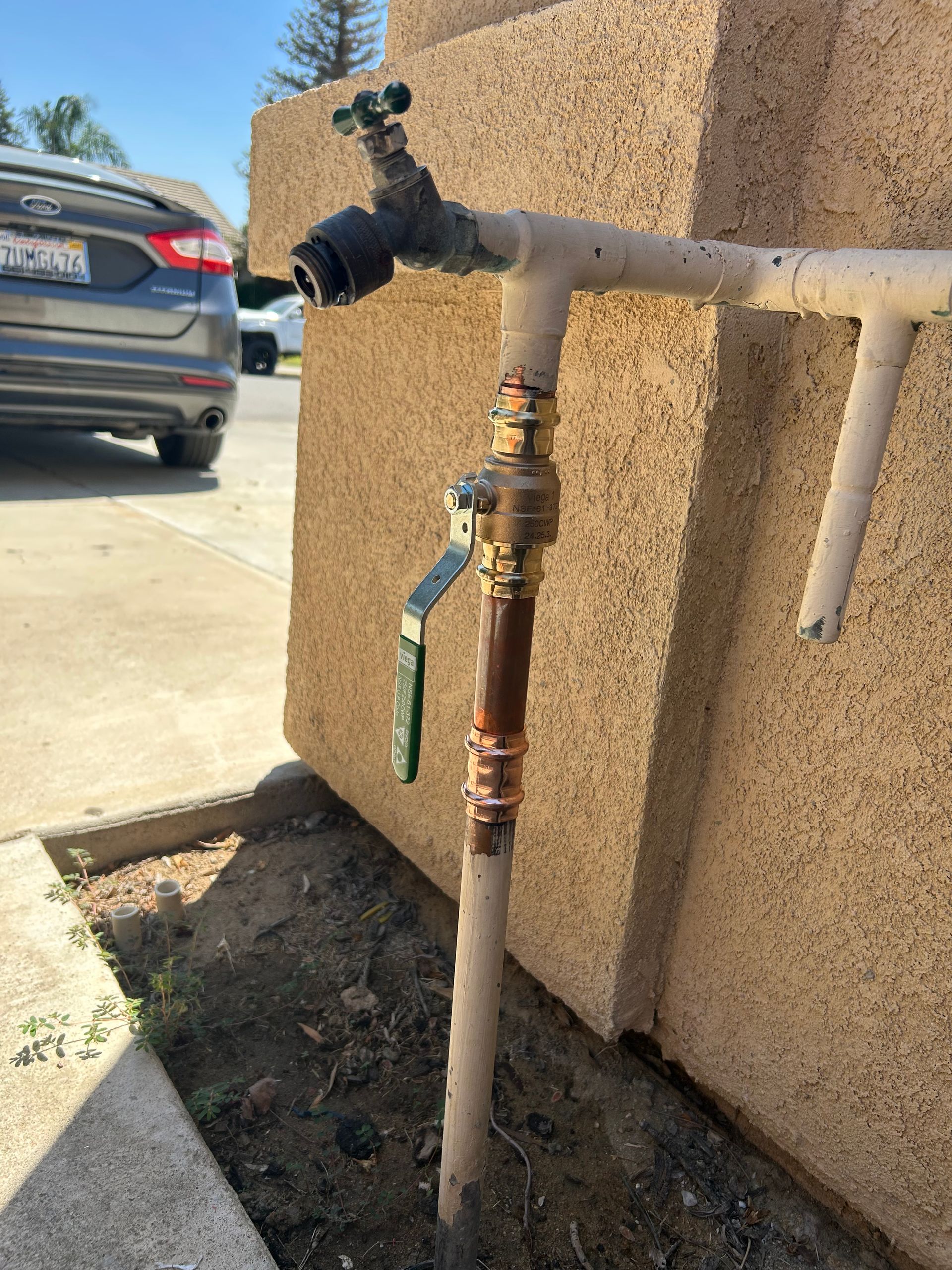 Outdoor water spigot with pipe and shutoff valve beside a stucco wall, near a driveway and parked car