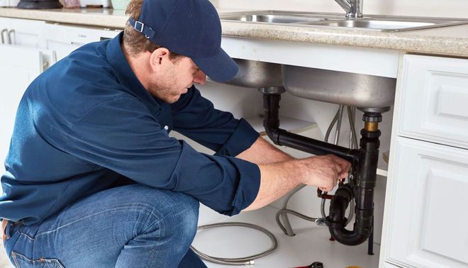 Plumber in blue uniform repairing pipes under a kitchen sink.