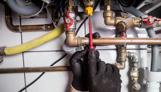 Gloved hands using a screwdriver to adjust plumbing valves and pipes under a sink