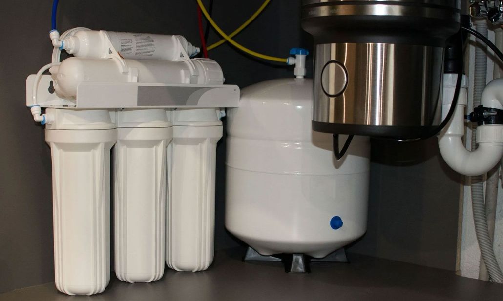 Man in blue overalls holding a tool beside a white water filtration unit in a kitchen