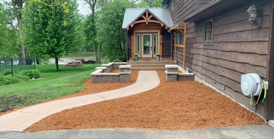 Path leads to a rustic house with a wooden porch. Mulch and lawn surround the pathway.