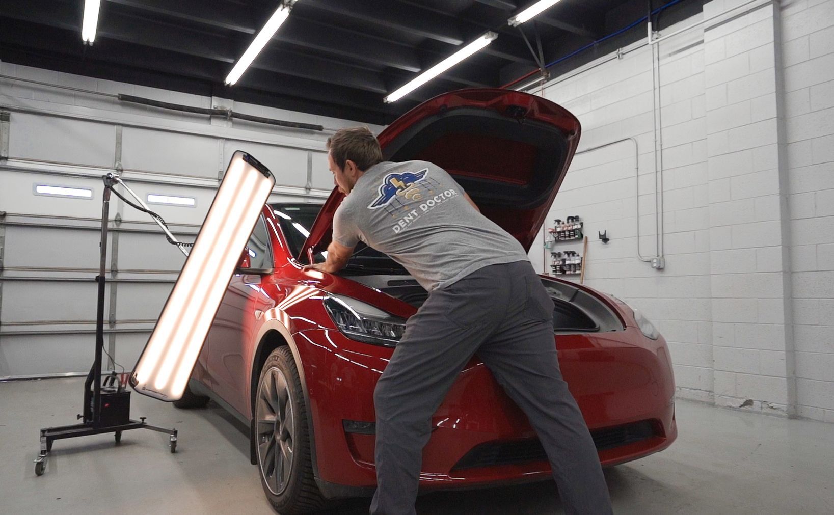 Man working on a red Tesla in a garage, hood open, with a light.