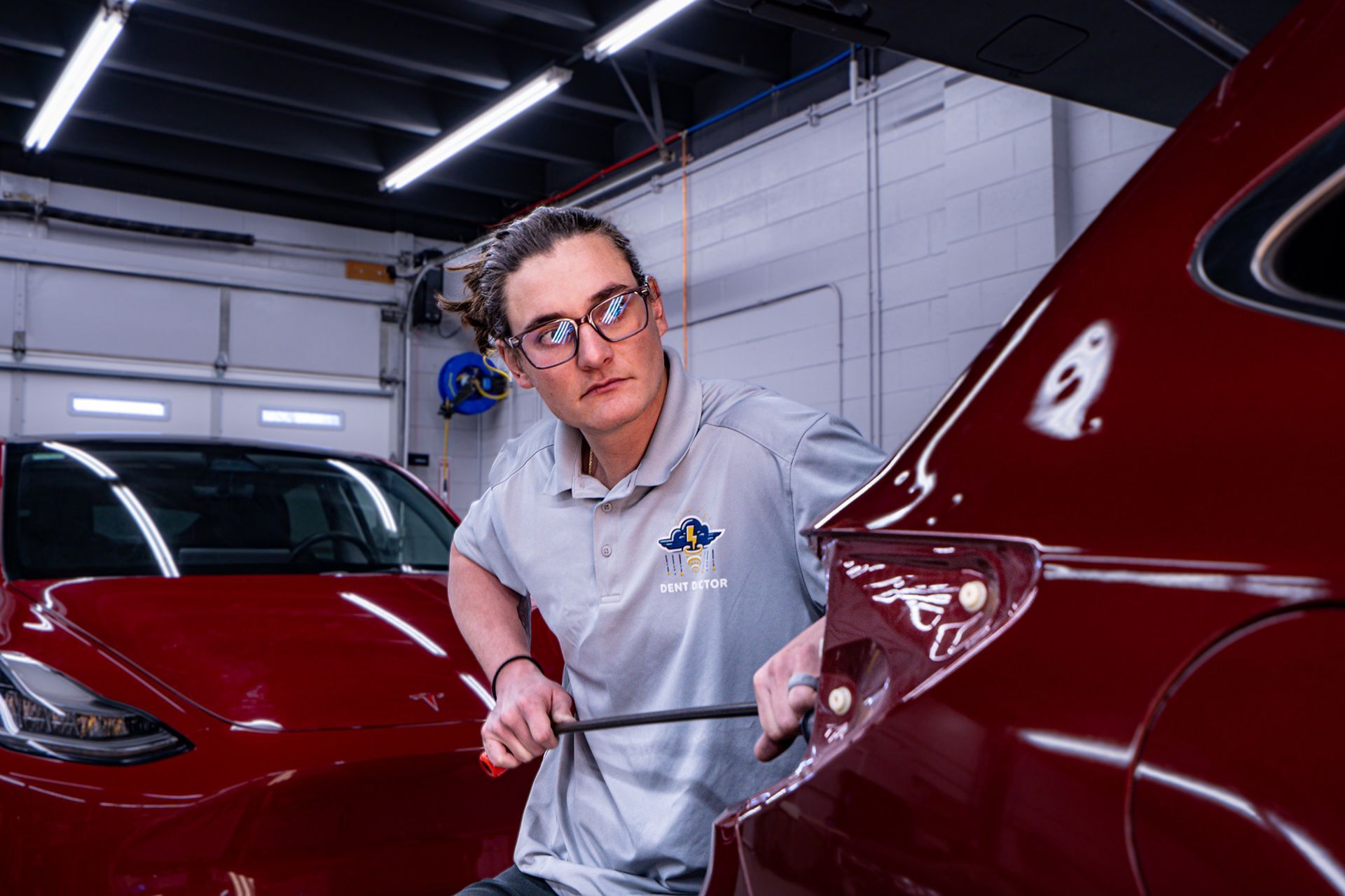 Mechanic working on a red car in a garage, looking focused. They wear glasses and a gray shirt.