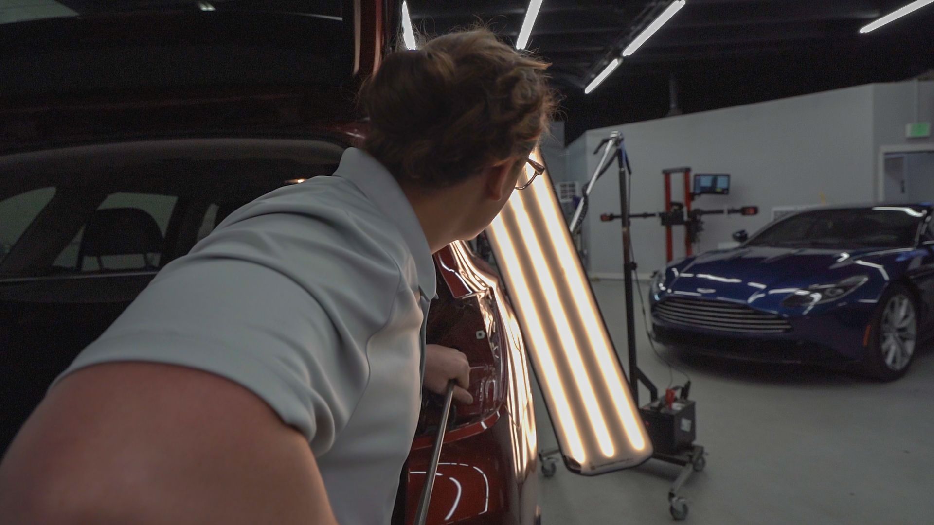 A person in a garage uses lights to inspect a dent on a red car's door. A blue car is in the background.