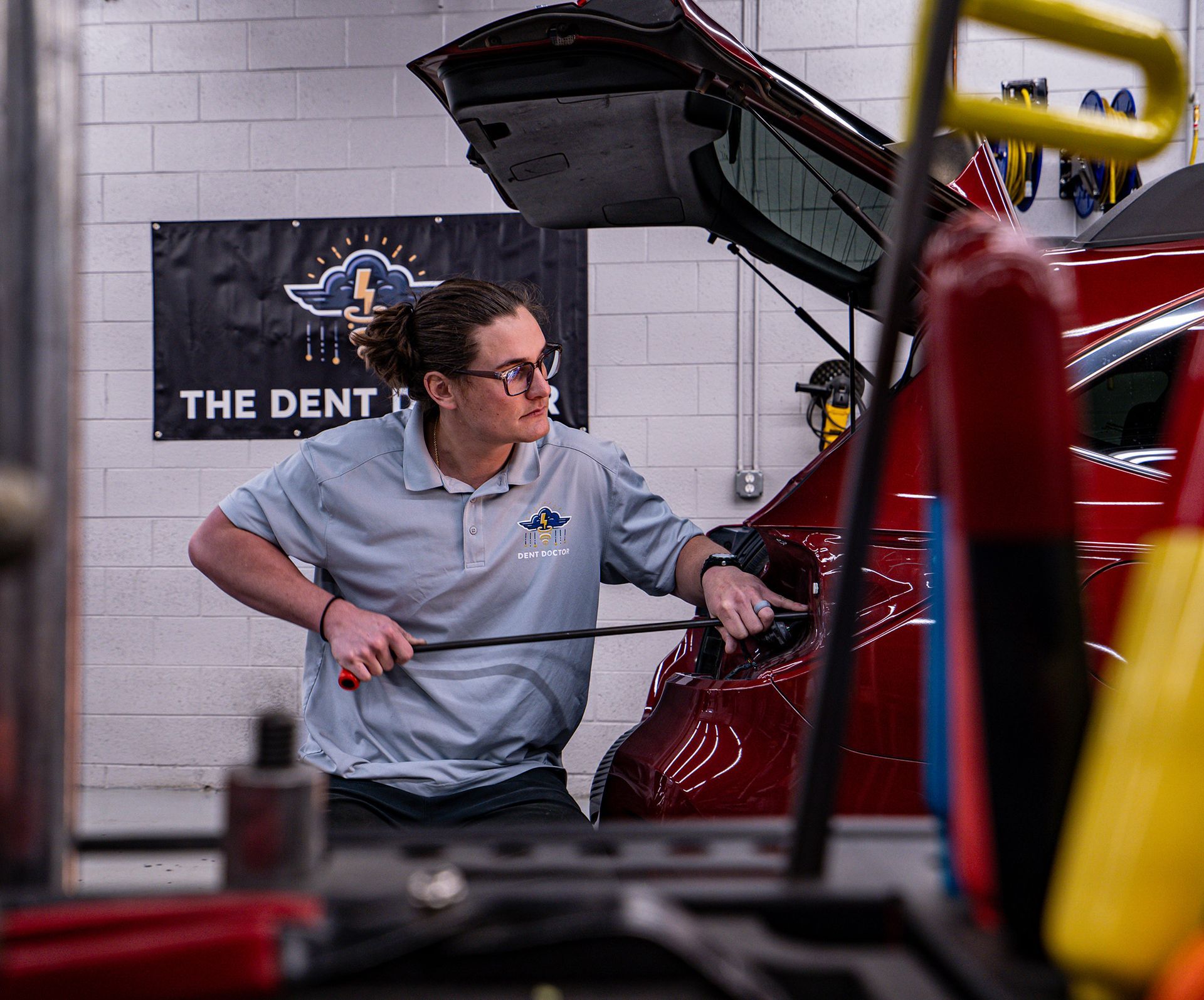 Man fixing a dent in a red car, using a tool inside the trunk. The setting is a repair shop.