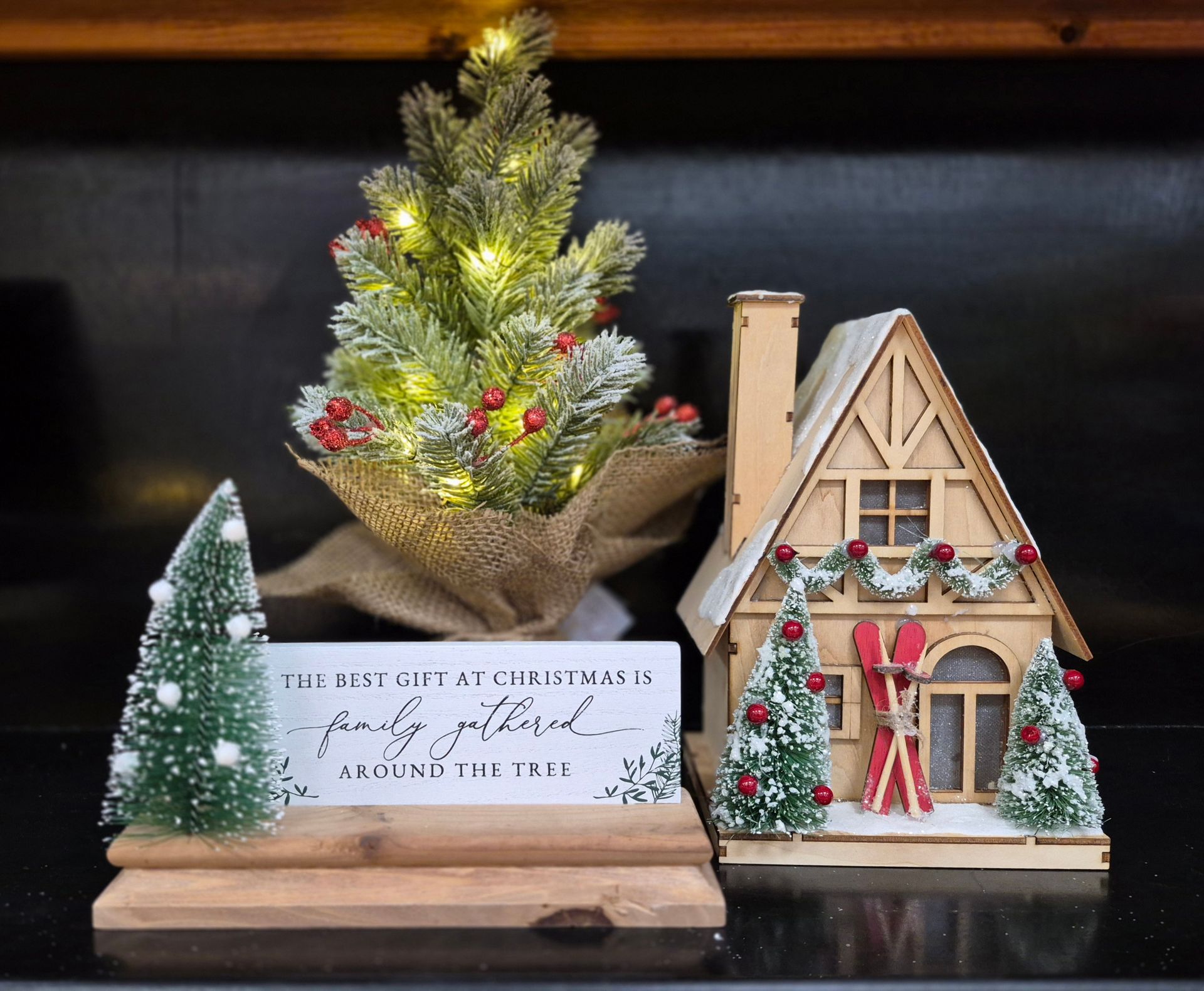 Christmas decor: mini tree with lights, rustic house with skis, sign, and smaller trees on a dark surface.